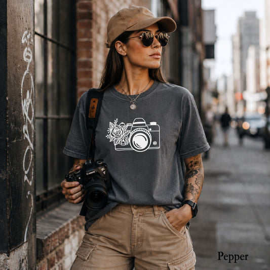A woman wearing a dark heather gray t-shirt with a white camera and floral graphic, paired with khaki cargo pants and a tan cap, standing on a city street.