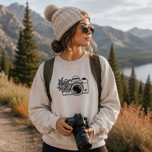 A woman outdoors wearing a cream knit beanie with a pom-pom, sunglasses, a backpack, and a cream sweatshirt with a black camera and floral graphic on the front. She is holding a DSLR camera in front of a scenic mountain and lake background.