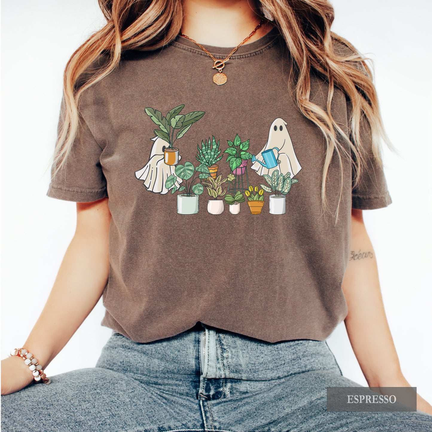 A woman wearing an espresso comfort colors t-shirt with ghosts tending to houseplants on it.