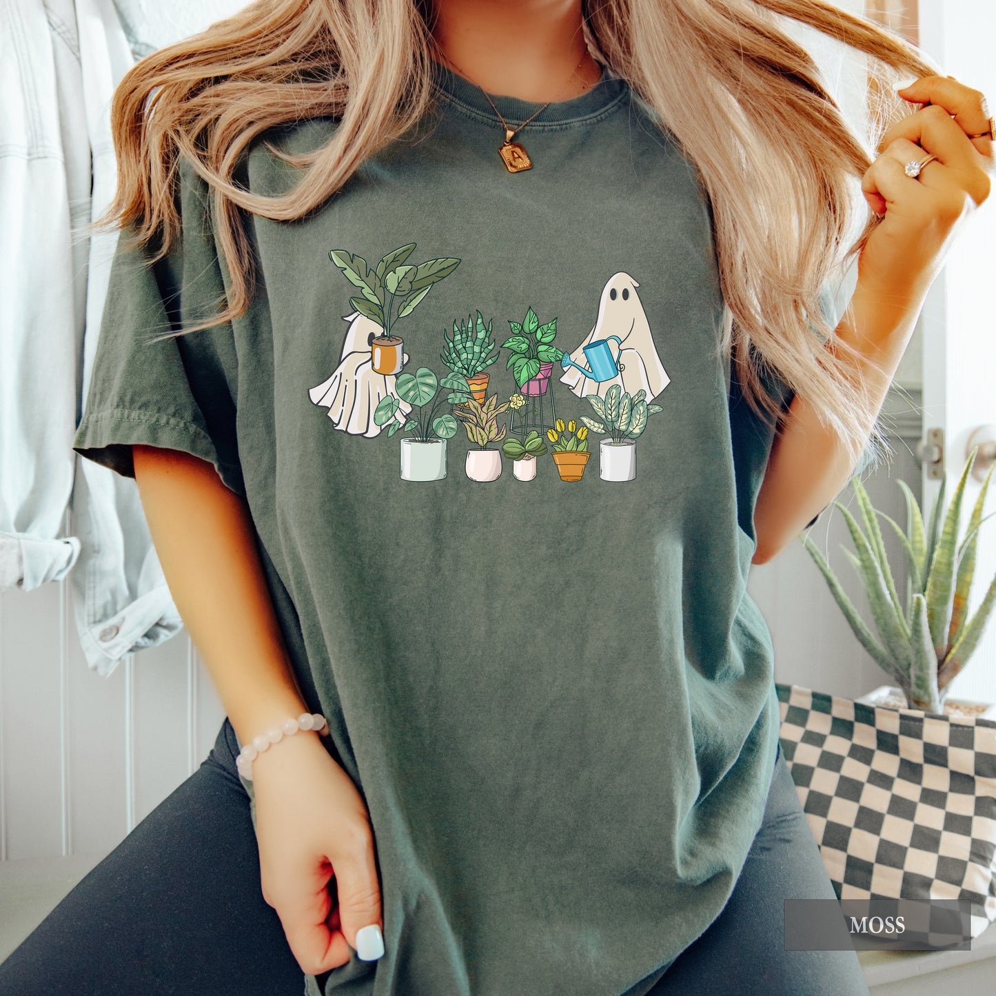 A woman wearing a moss comfort colors t-shirt with ghosts tending to houseplants on it.
