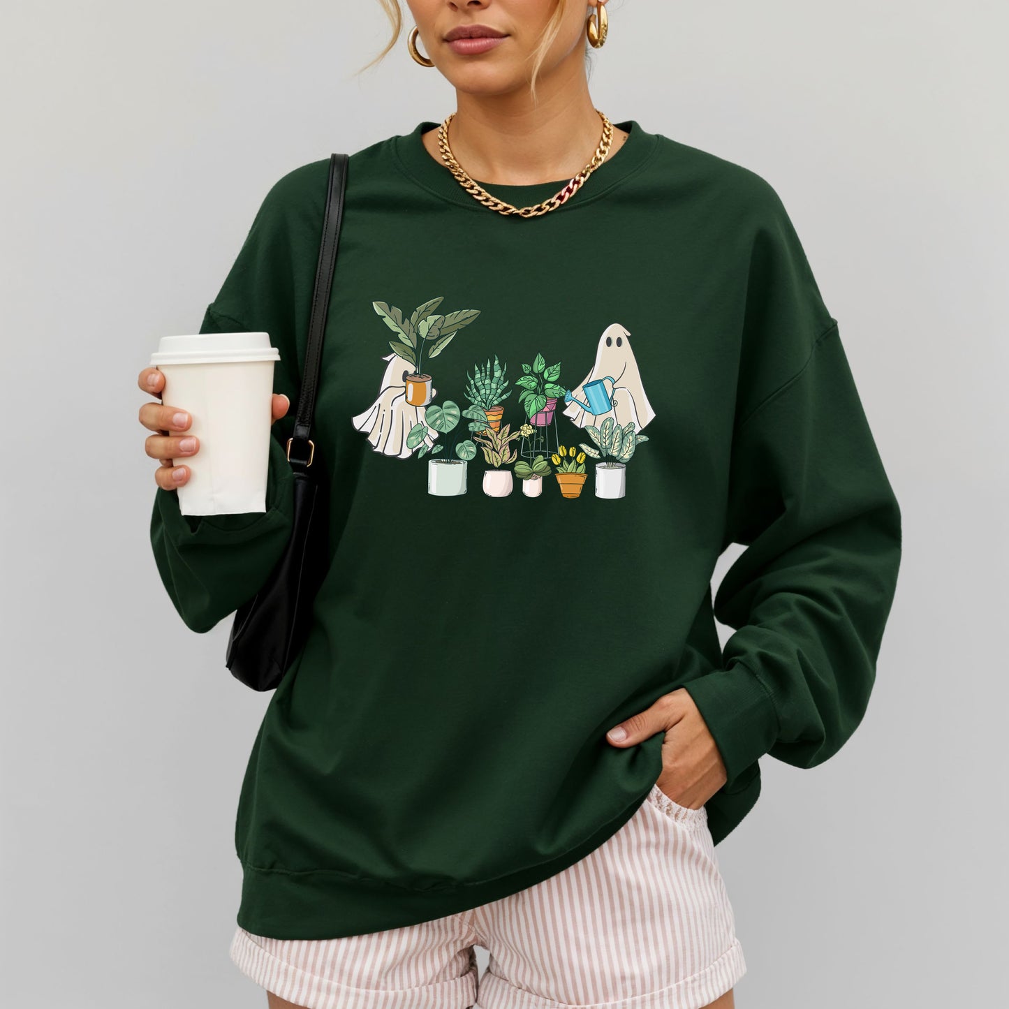 Woman wearing a forest green gildan sweatshirt with an image of ghosts tending houseplants on it.