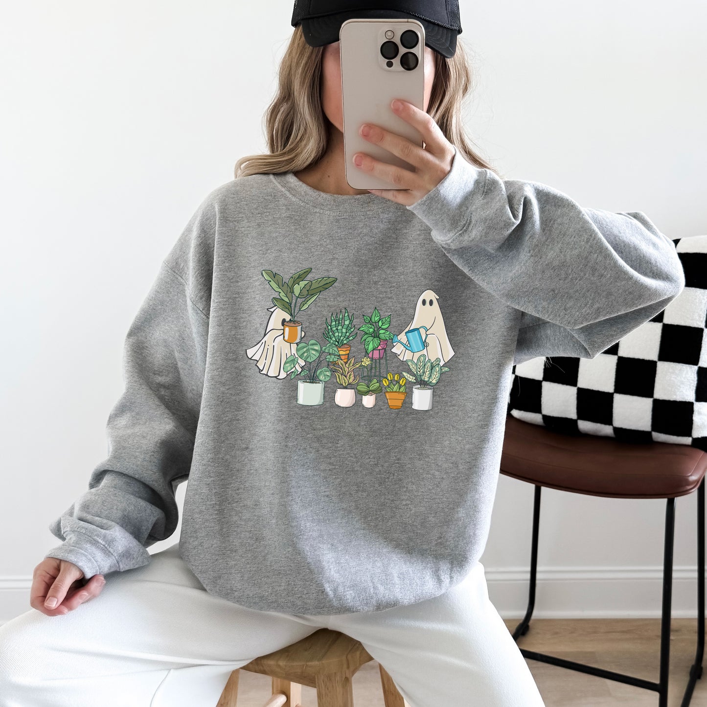 Woman wearing a sport grey gildan sweatshirt with an image of ghosts tending houseplants on it.