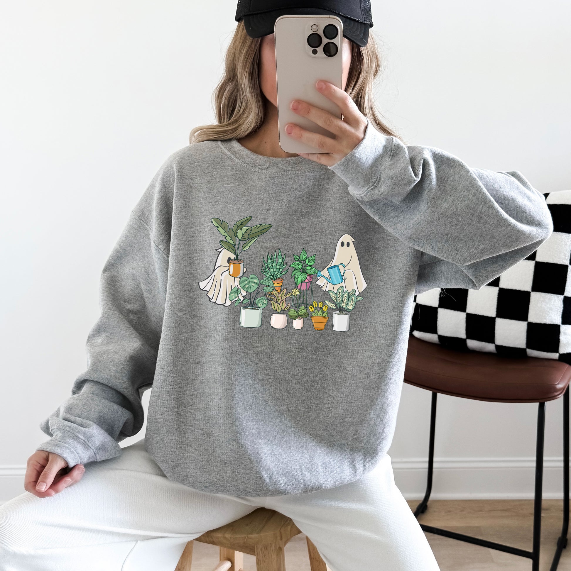 Woman wearing a sport grey gildan sweatshirt with an image of ghosts tending houseplants on it.