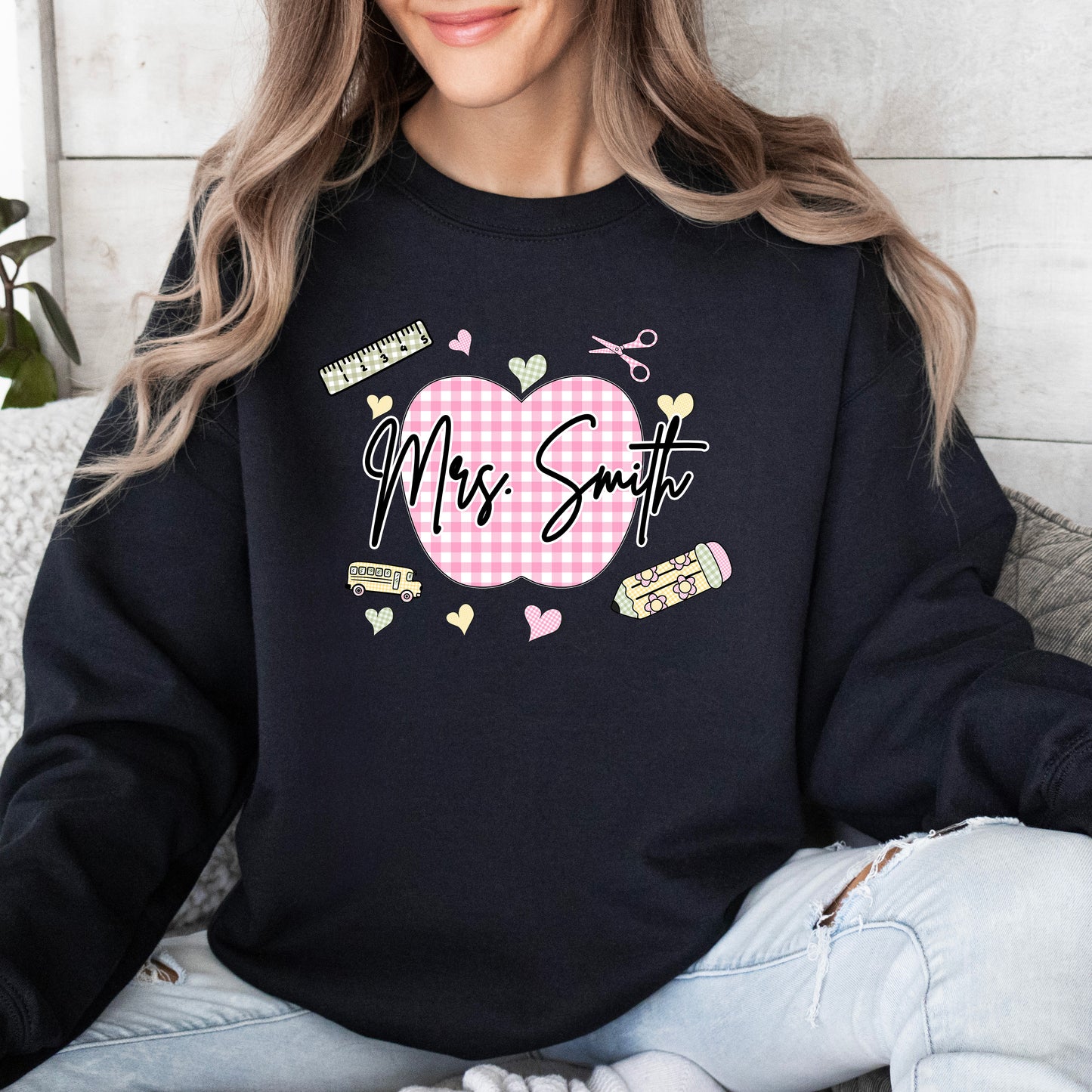 Teacher wearing a black sweatshirt with a graphic design that's personalized with her name, a pink gingham apple, and gingham school supplies.