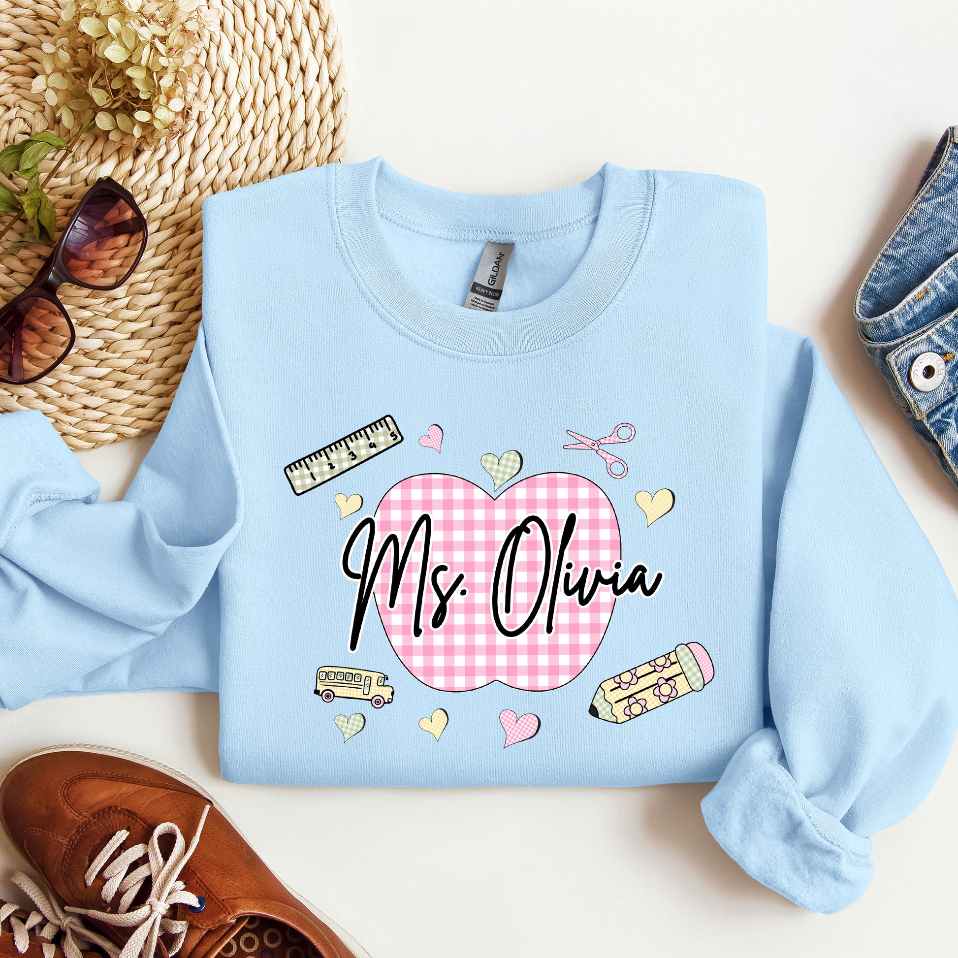 Teacher wearing a light blue sweatshirt with a graphic design that's personalized with her name, a pink gingham apple, and gingham school supplies.