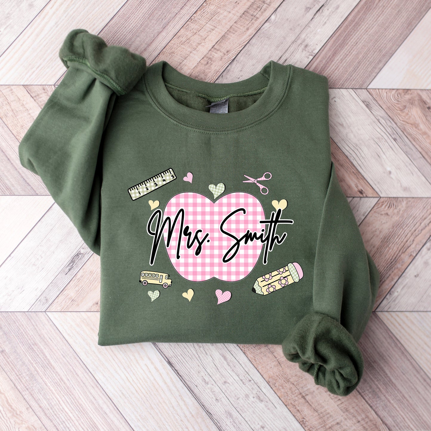 Teacher wearing a military green sweatshirt with a graphic design that's personalized with her name, a pink gingham apple, and gingham school supplies.