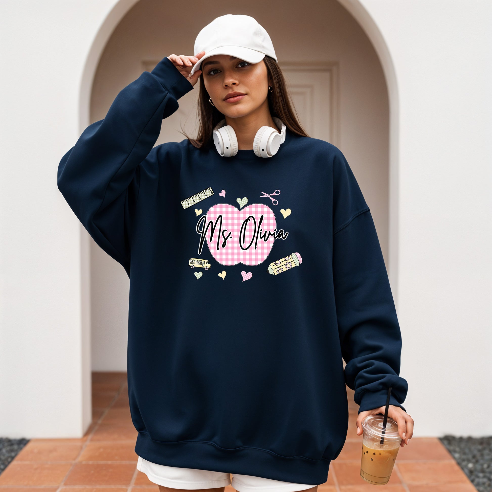 Teacher wearing a navy sweatshirt with a graphic design that's personalized with her name, a pink gingham apple, and gingham school supplies, holding a coffee cup outdoors.
