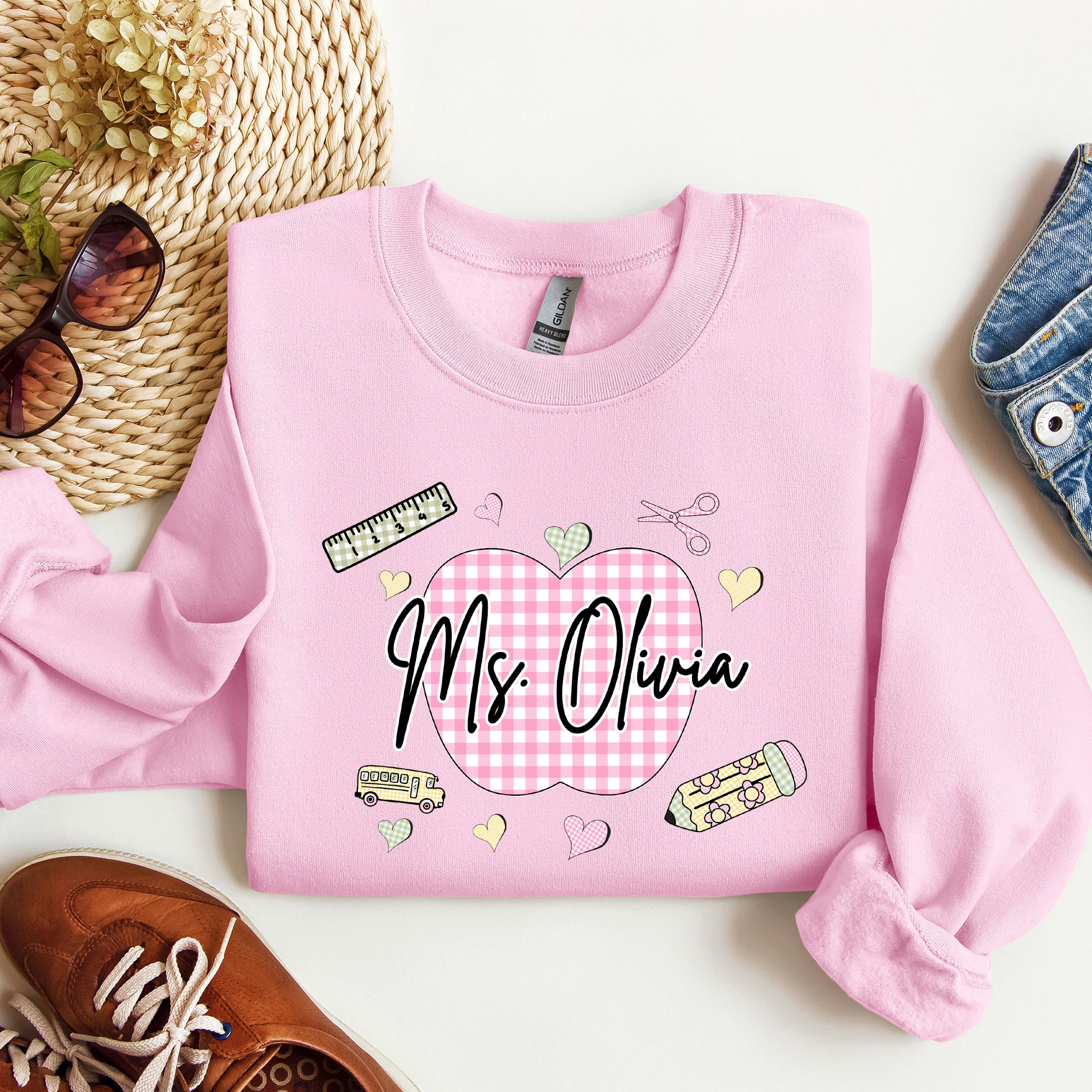 Teacher wearing a light pink sweatshirt with a graphic design that's personalized with her name, a pink gingham apple, and gingham school supplies.