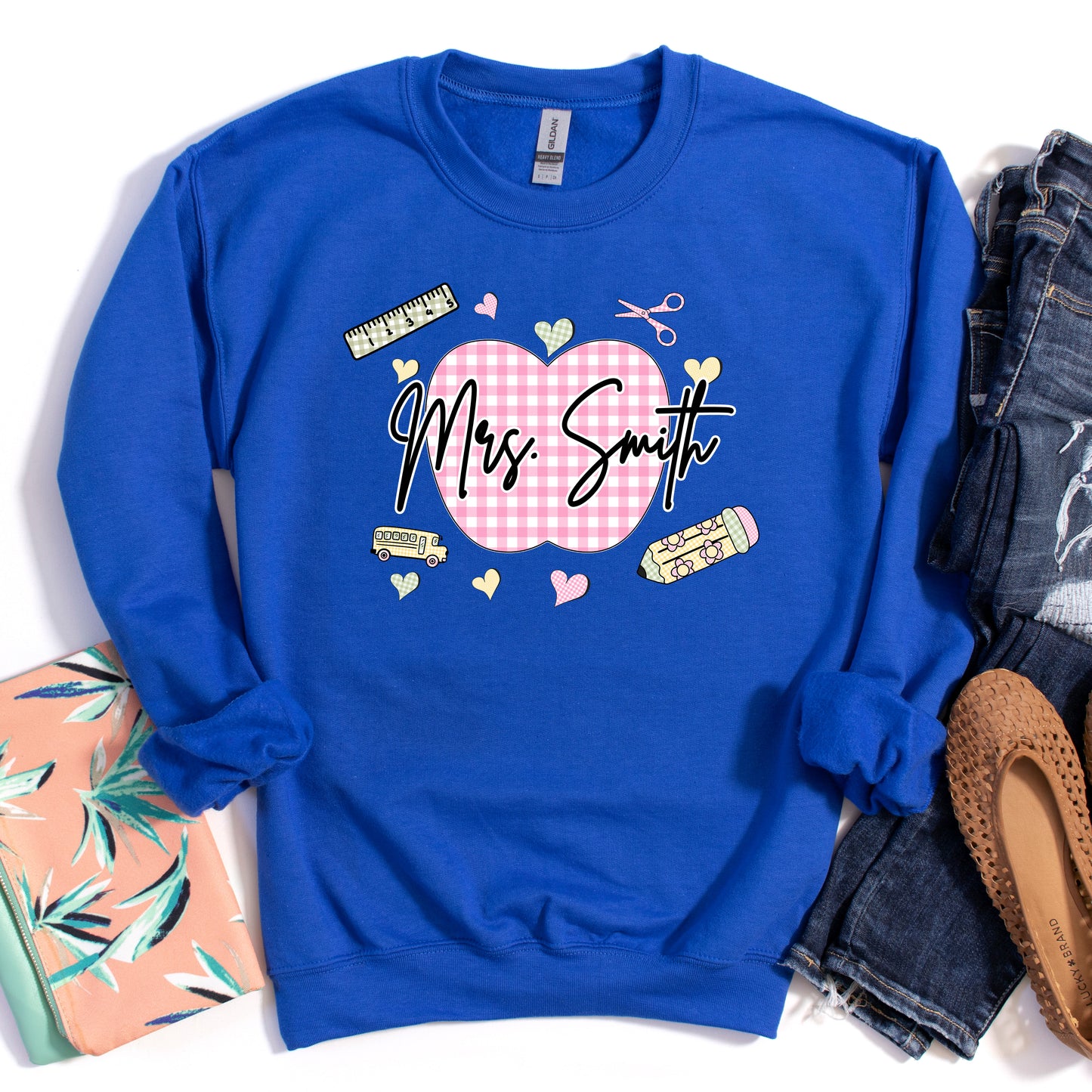 Teacher wearing a royal blue sweatshirt with a graphic design that's personalized with her name, a pink gingham apple, and gingham school supplies.