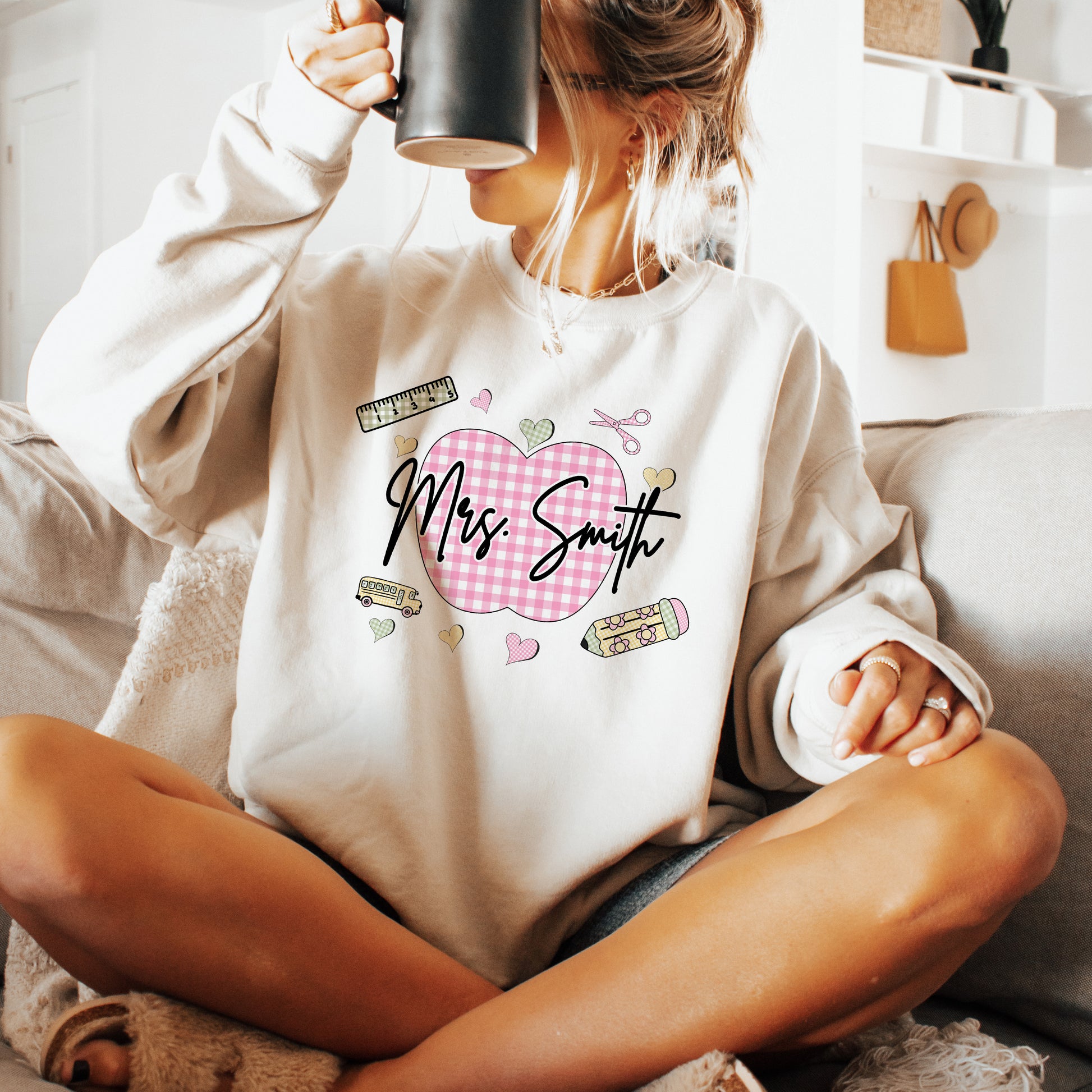 Teacher wearing a sand sweatshirt with a graphic design that's personalized with her name, a pink gingham apple, and gingham school supplies.