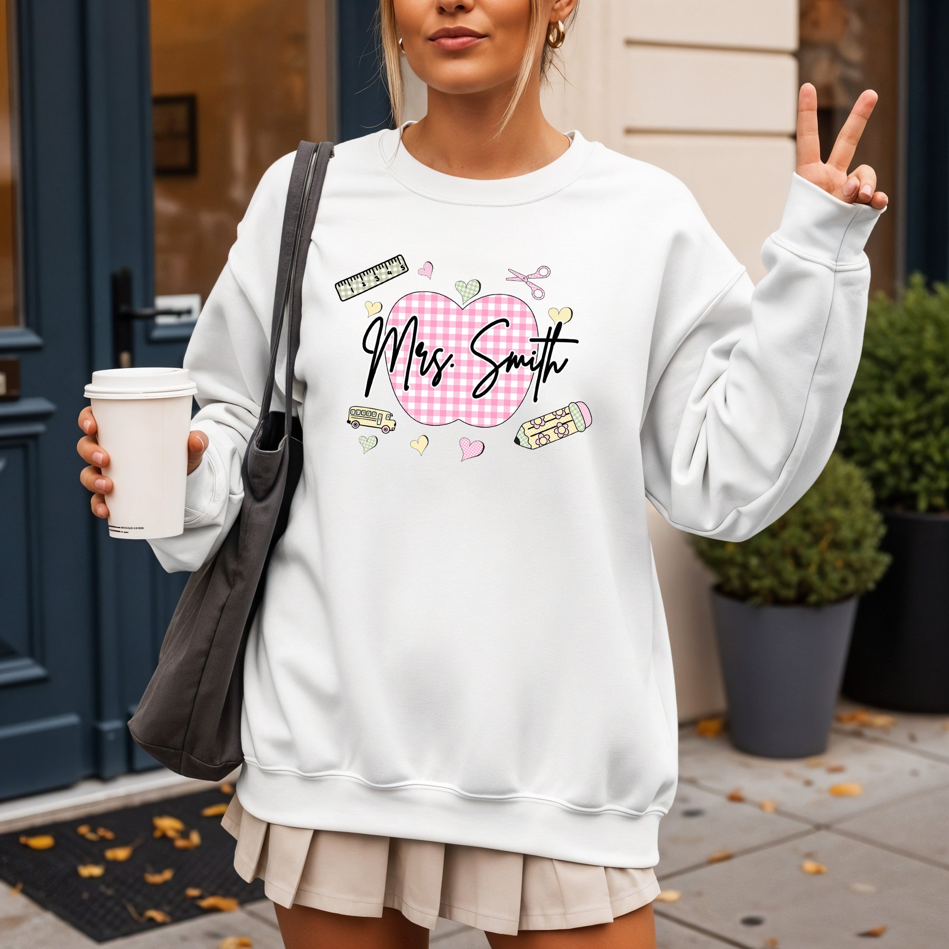 Teacher wearing a white sweatshirt with a graphic design that's personalized with her name, a pink gingham apple, and gingham school supplies, holding a coffee cup outdoors.