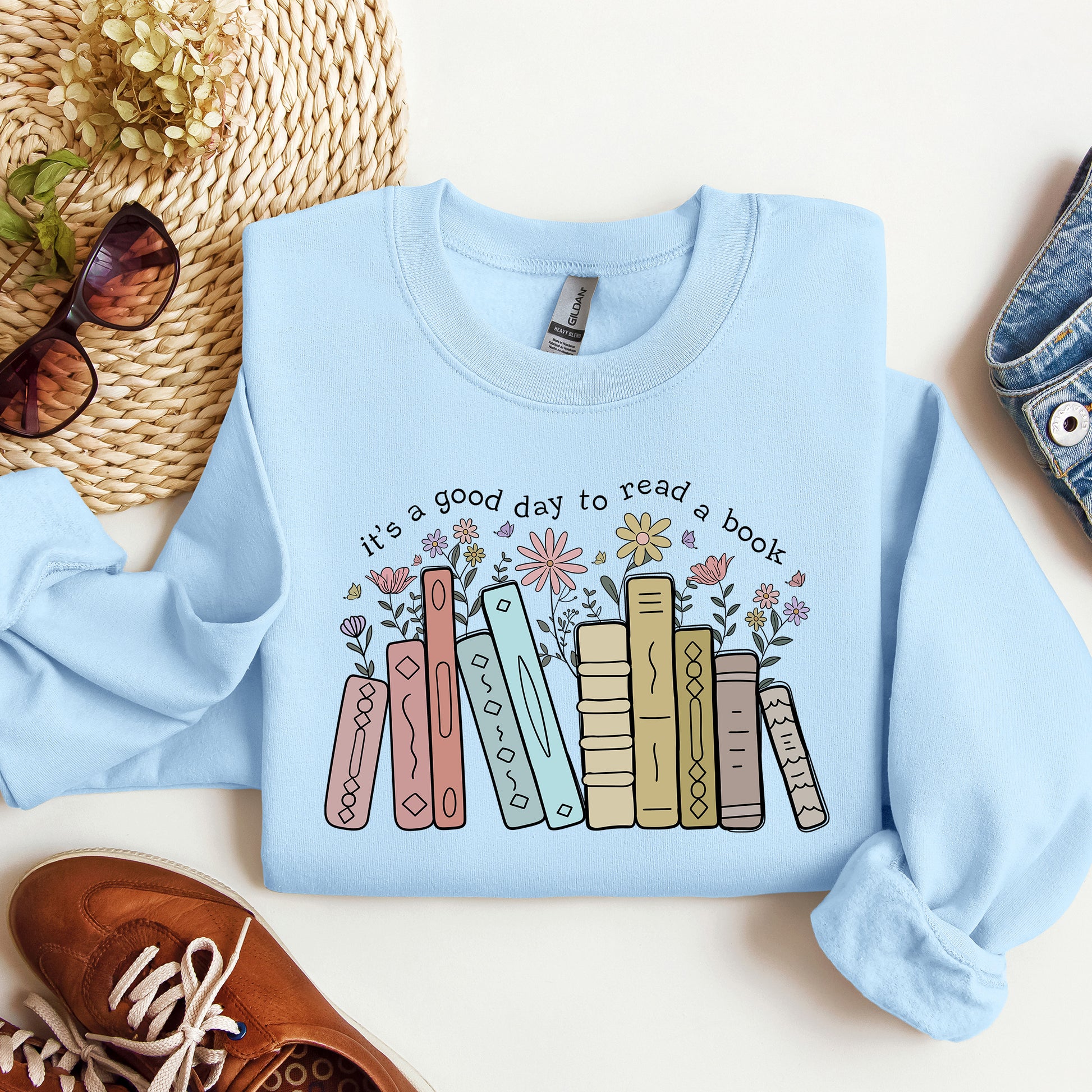 Light blue sweatshirt with book design and text 'it's a good day to read a book' on a white surface with accessories.