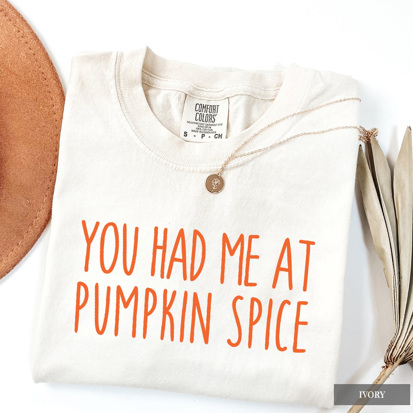 A folded ivory crewneck t-shirt with orange text reading 'YOU HAD ME AT PUMPKIN SPICE' on the front, styled with a necklace and autumn-themed props on a white background.