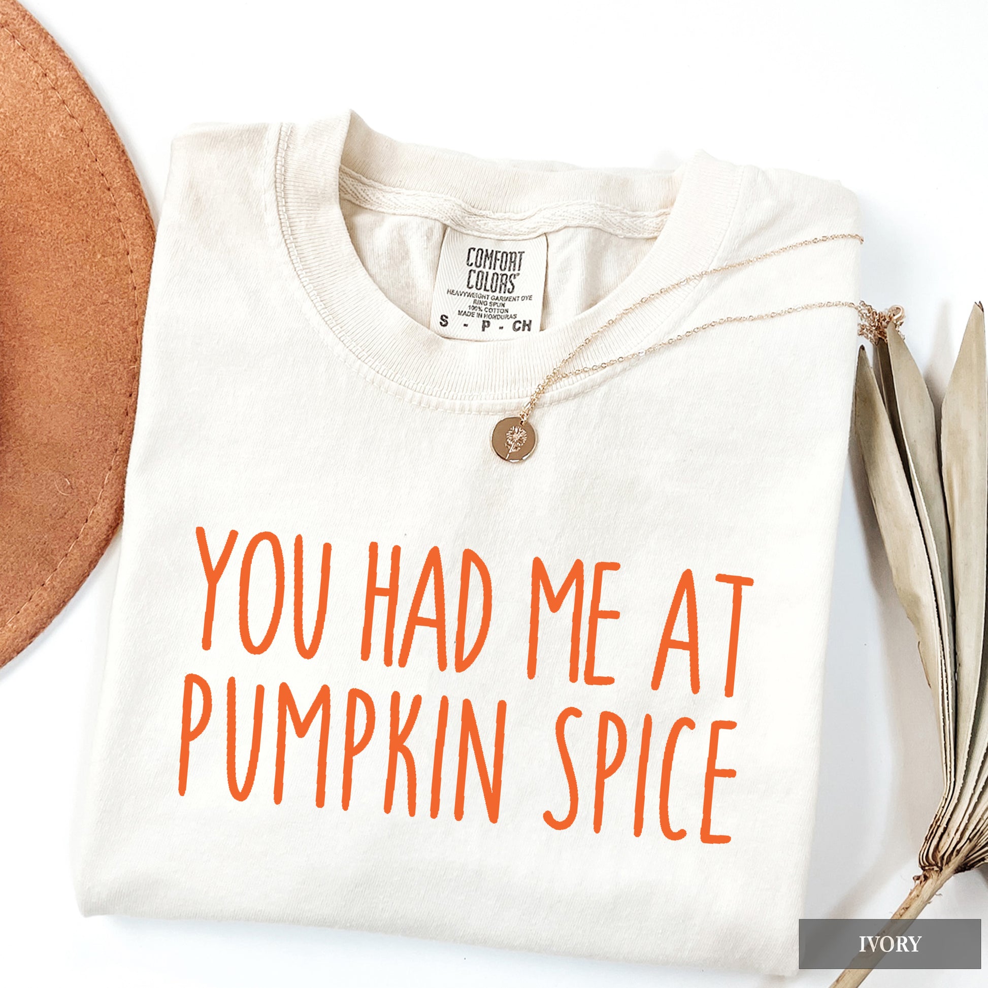 A folded ivory crewneck t-shirt with orange text reading 'YOU HAD ME AT PUMPKIN SPICE' on the front, styled with a necklace and autumn-themed props on a white background.