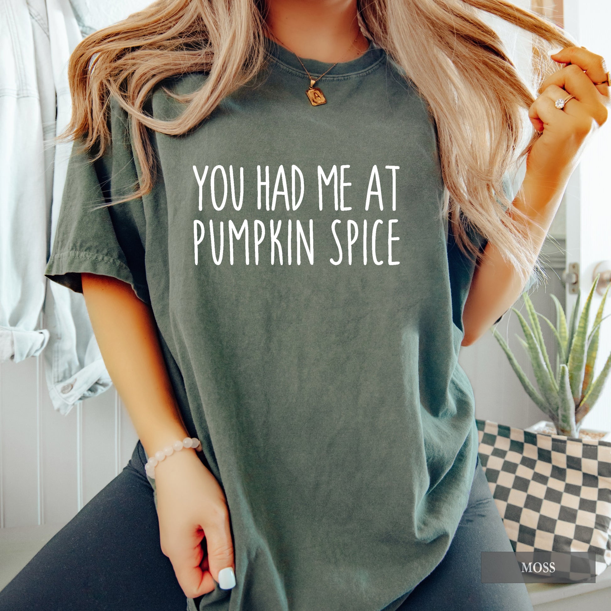 A woman wearing an oversized moss green t-shirt with the phrase 'YOU HAD ME AT PUMPKIN SPICE' printed in white across the chest. The photo is taken in a lifestyle setting with a window, plant, and clothing rack in the background.