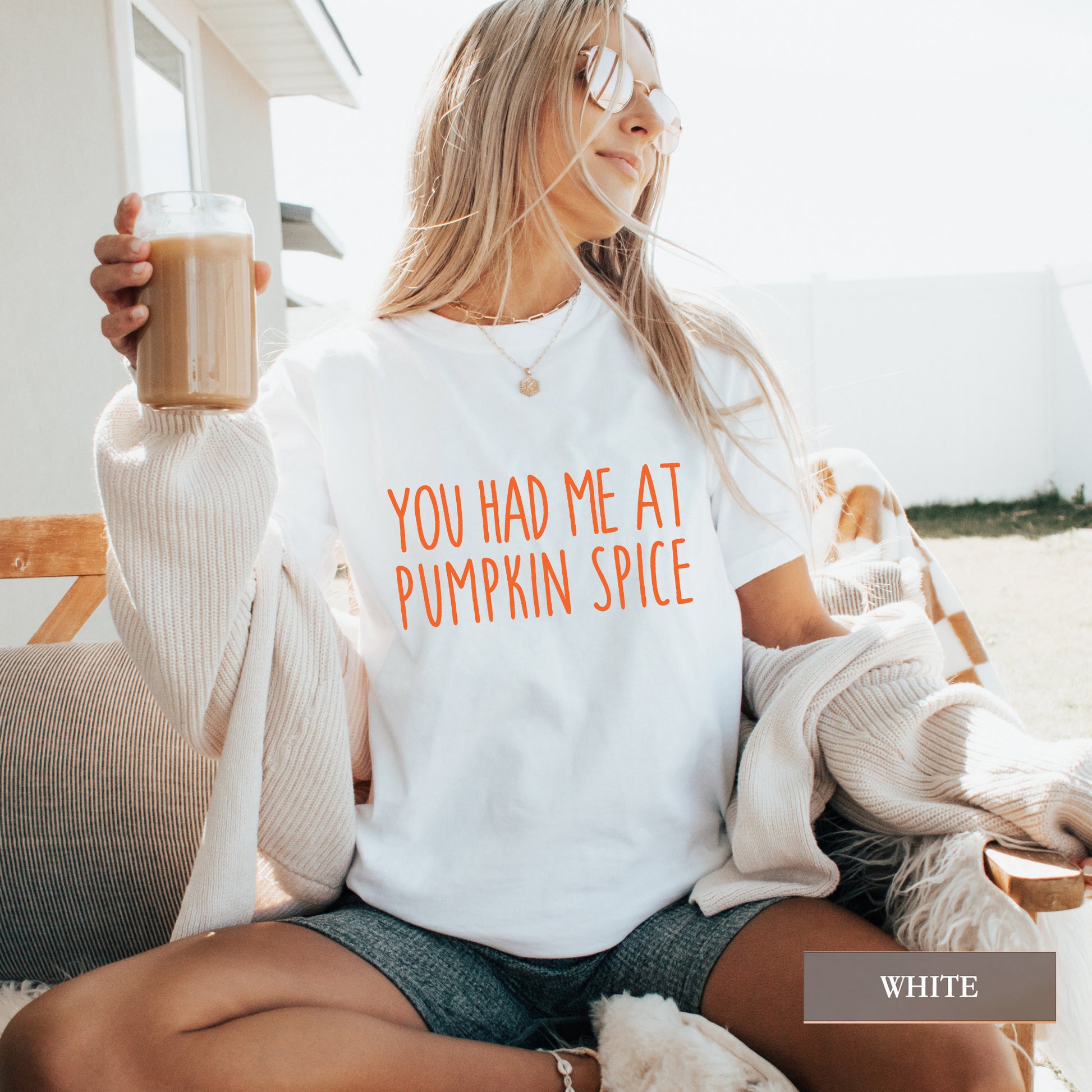 A woman sitting outdoors wearing a white graphic T-shirt with orange text that reads 'YOU HAD ME AT PUMPKIN SPICE'. She is holding a drink and has a cream knit cardigan draped off her shoulders.