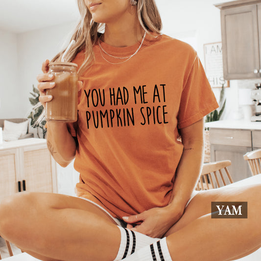 A woman wearing a burnt orange graphic t-shirt with the text 'YOU HAD ME AT PUMPKIN SPICE' in black. She is holding an iced coffee in a bright kitchen setting, wearing white socks with black stripes.