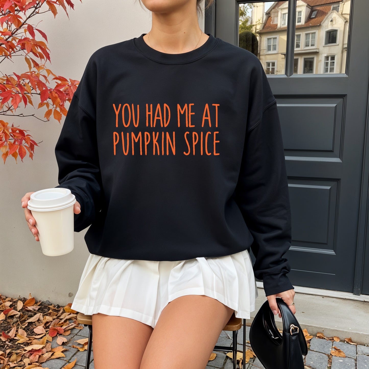 A woman wearing a black crewneck sweatshirt with orange text that reads 'YOU HAD ME AT PUMPKIN SPICE', paired with white shorts, holding a coffee cup and a black handbag in an outdoor setting.