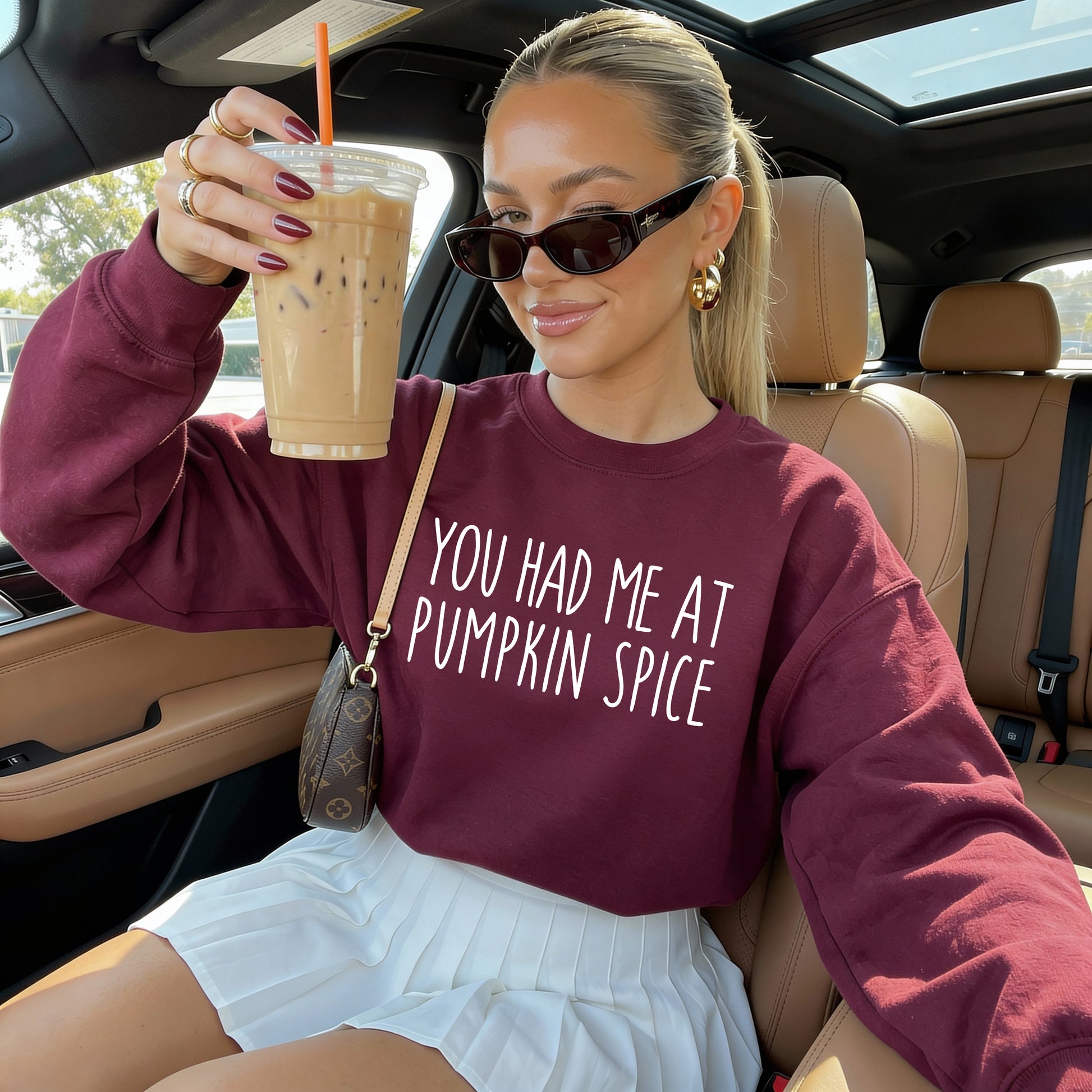 A woman sitting in a car holds up an iced coffee while wearing a maroon sweatshirt with the white text 'YOU HAD ME AT PUMPKIN SPICE'.