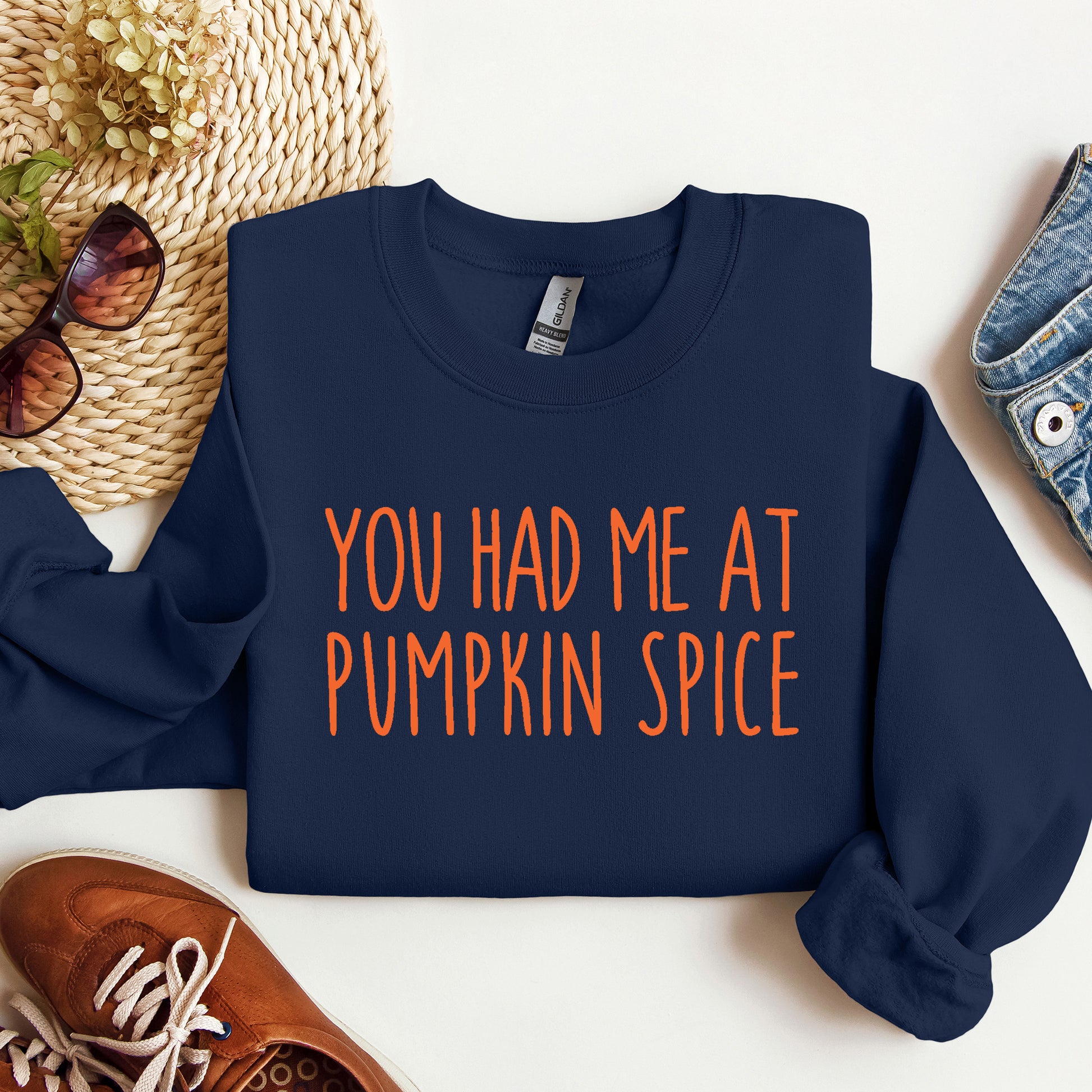 A navy blue sweatshirt with the phrase 'YOU HAD ME AT PUMPKIN SPICE' printed in orange across the front, styled in a flat lay with sunglasses, a woven hat, jeans, and brown shoes on a white background.