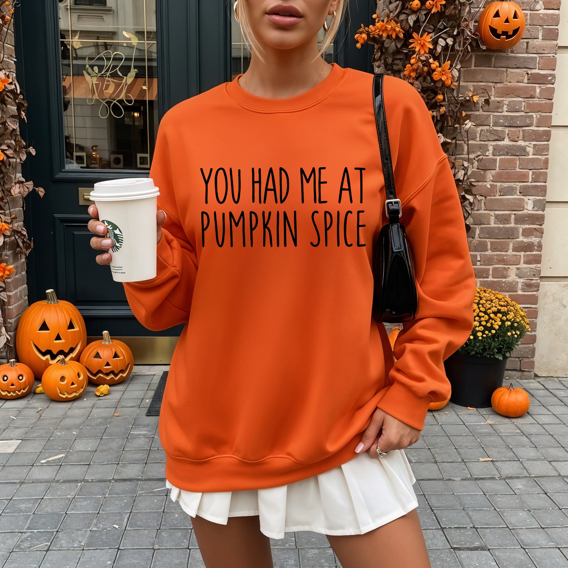 A woman wearing an oversized bright orange sweatshirt with the text 'YOU HAD ME AT PUMPKIN SPICE' in black lettering. She is holding a white Starbucks coffee cup and standing outdoors in front of a brick door decorated with pumpkins.
