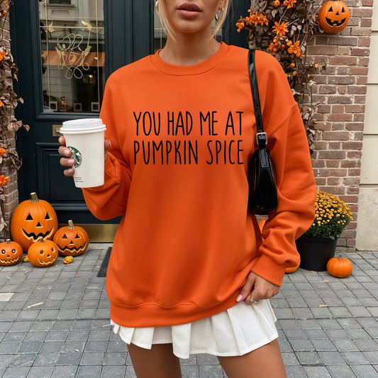 A woman wearing an oversized bright orange sweatshirt with the text 'YOU HAD ME AT PUMPKIN SPICE' in black lettering. She is holding a white Starbucks coffee cup and standing outdoors in front of a brick door decorated with pumpkins.