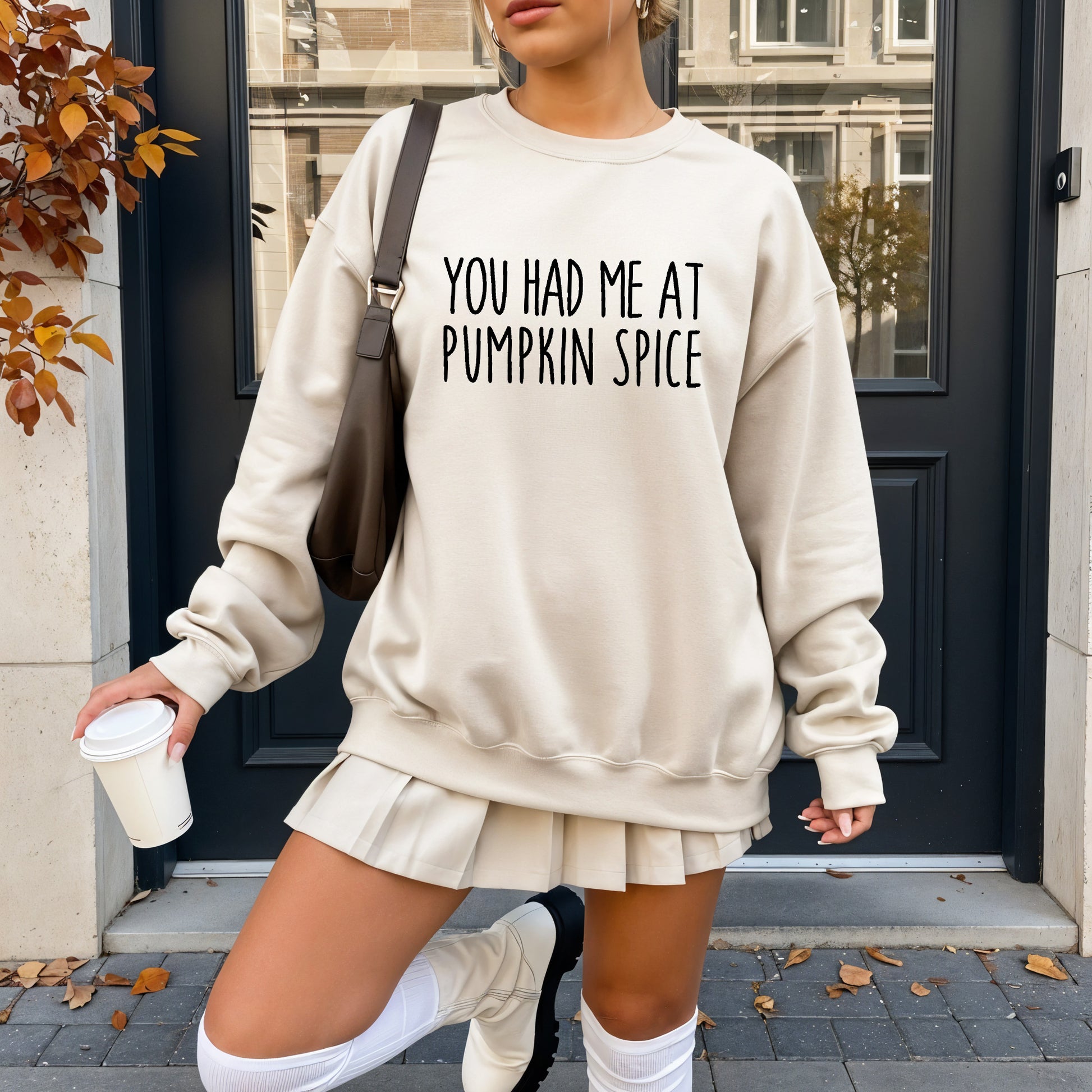 A woman wearing a cream-colored sweatshirt with the text 'YOU HAD ME AT PUMPKIN SPICE' in black capital letters, paired with a matching pleated mini skirt, white knee-high socks, and a dark brown shoulder bag, standing outdoors in front of a dark door.