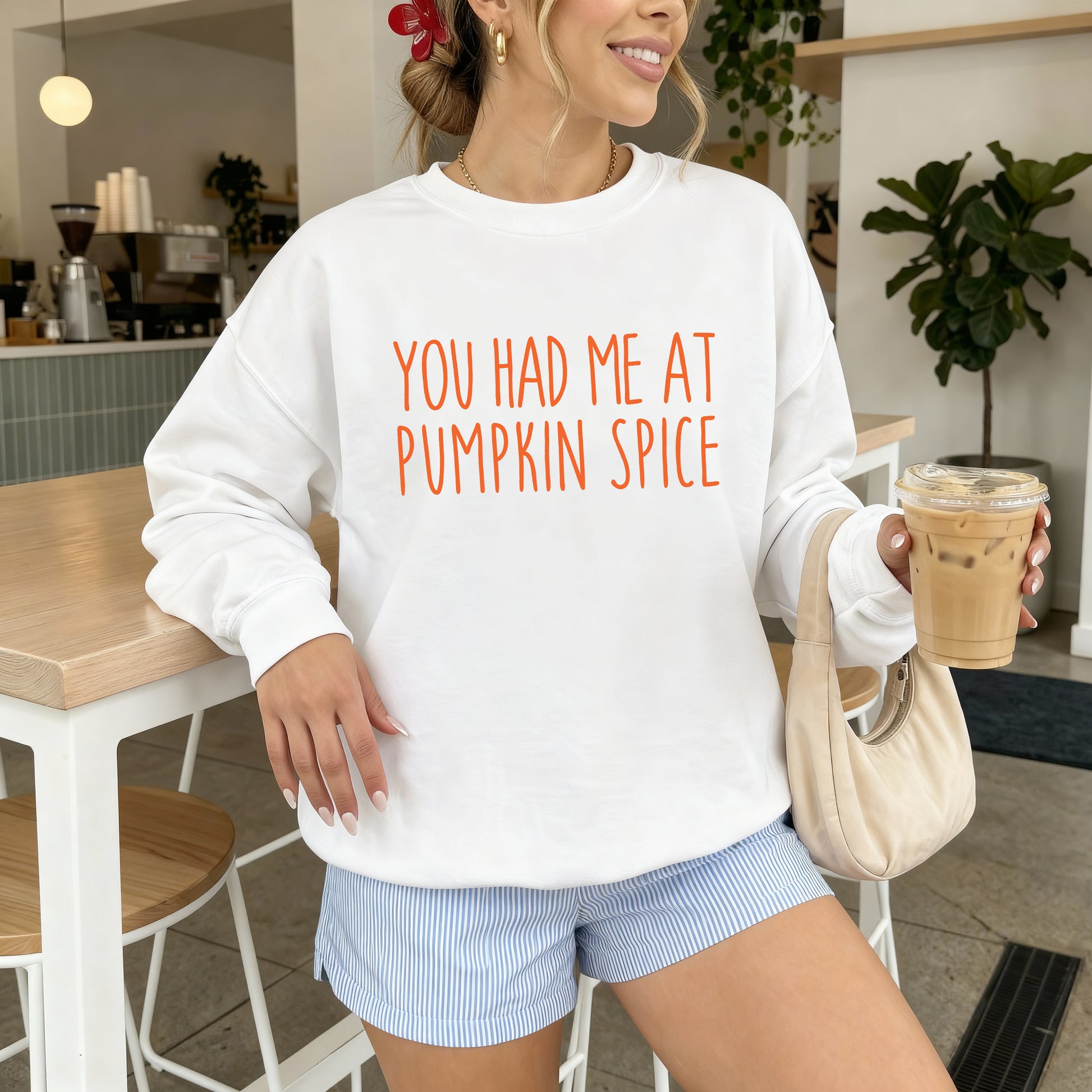 A woman wearing a white crewneck sweatshirt with orange text that reads 'YOU HAD ME AT PUMPKIN SPICE', paired with light blue and white striped shorts, holding an iced coffee in a cafe setting.