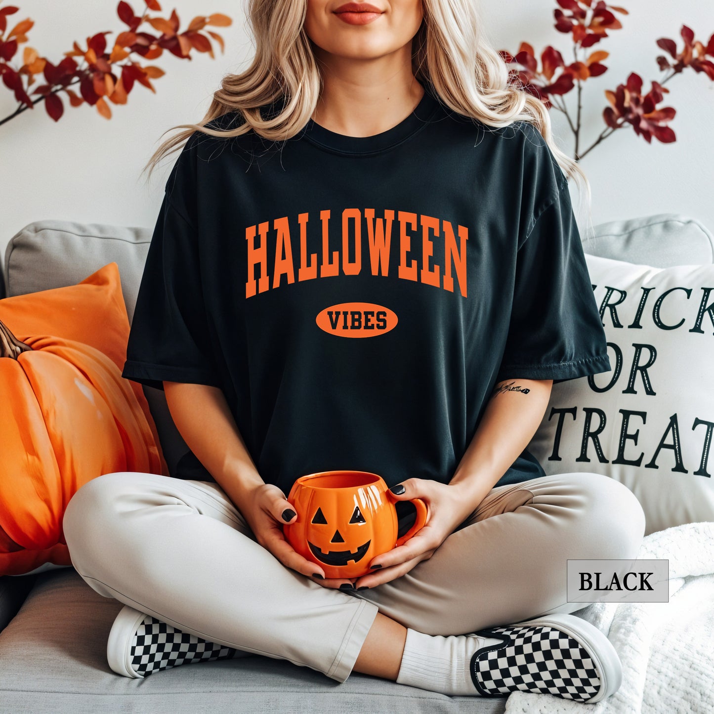 A woman sits cross-legged on a couch wearing a black graphic T-shirt with orange text reading 'HALLOWEEN VIBES' and holding an orange jack-o'-lantern mug. The scene includes pumpkins, pillows, and fall leaves in the background, with a small 'BLACK' label in the lower right corner.
