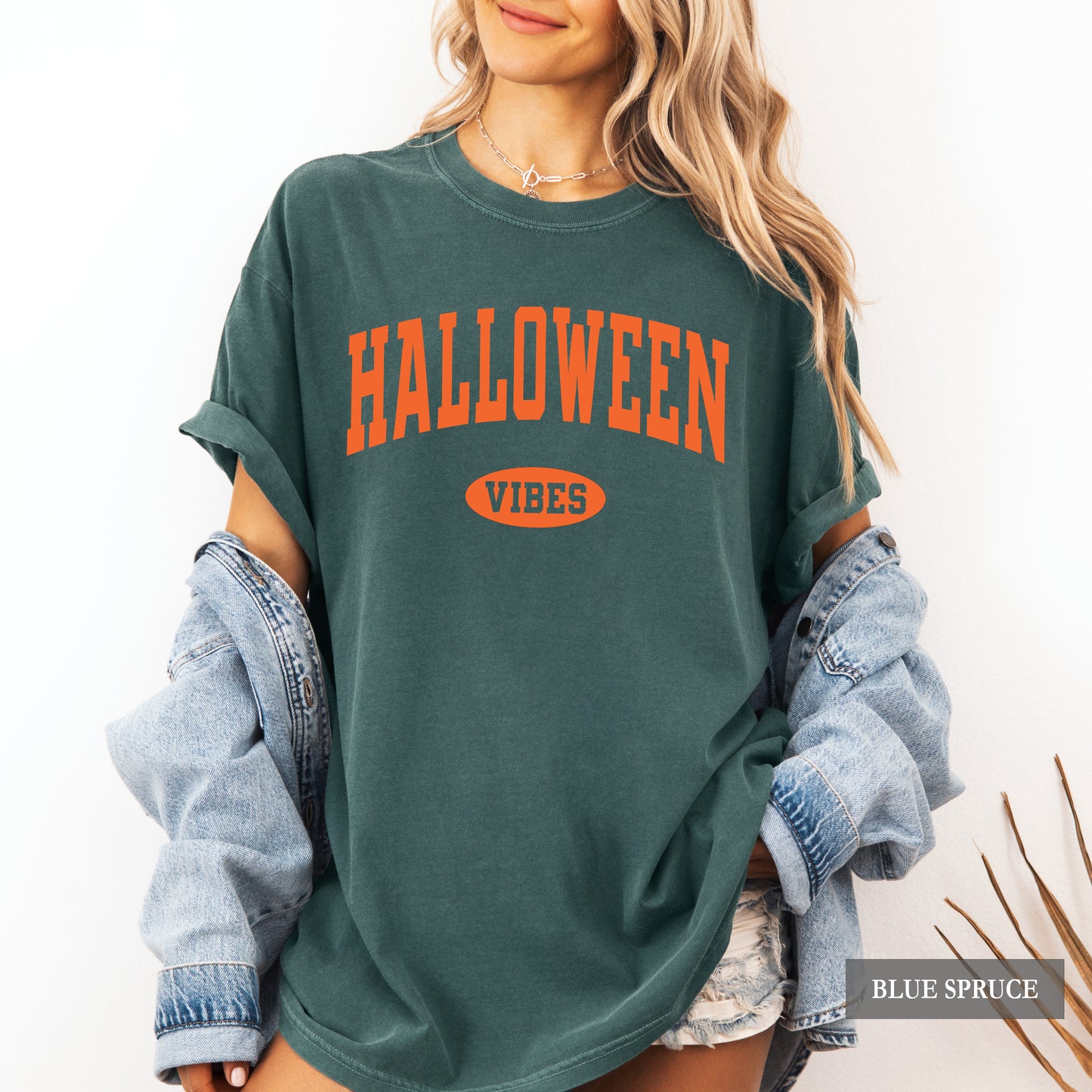 A woman wearing an oversized dark teal-green graphic t-shirt with 'HALLOWEEN VIBES' printed in orange on the front, styled with rolled-up light-wash denim sleeves.