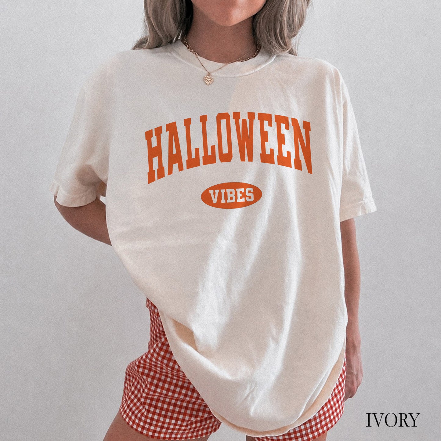 A person wearing an oversized off-white graphic T-shirt with orange text reading 'HALLOWEEN VIBES' on the front, paired with red-and-white gingham shorts against a plain light gray background.