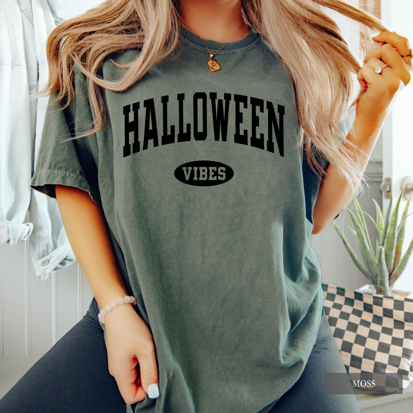 A woman wearing a moss green graphic t-shirt with 'HALLOWEEN VIBES' printed in black on the front, posed in a bright indoor setting.