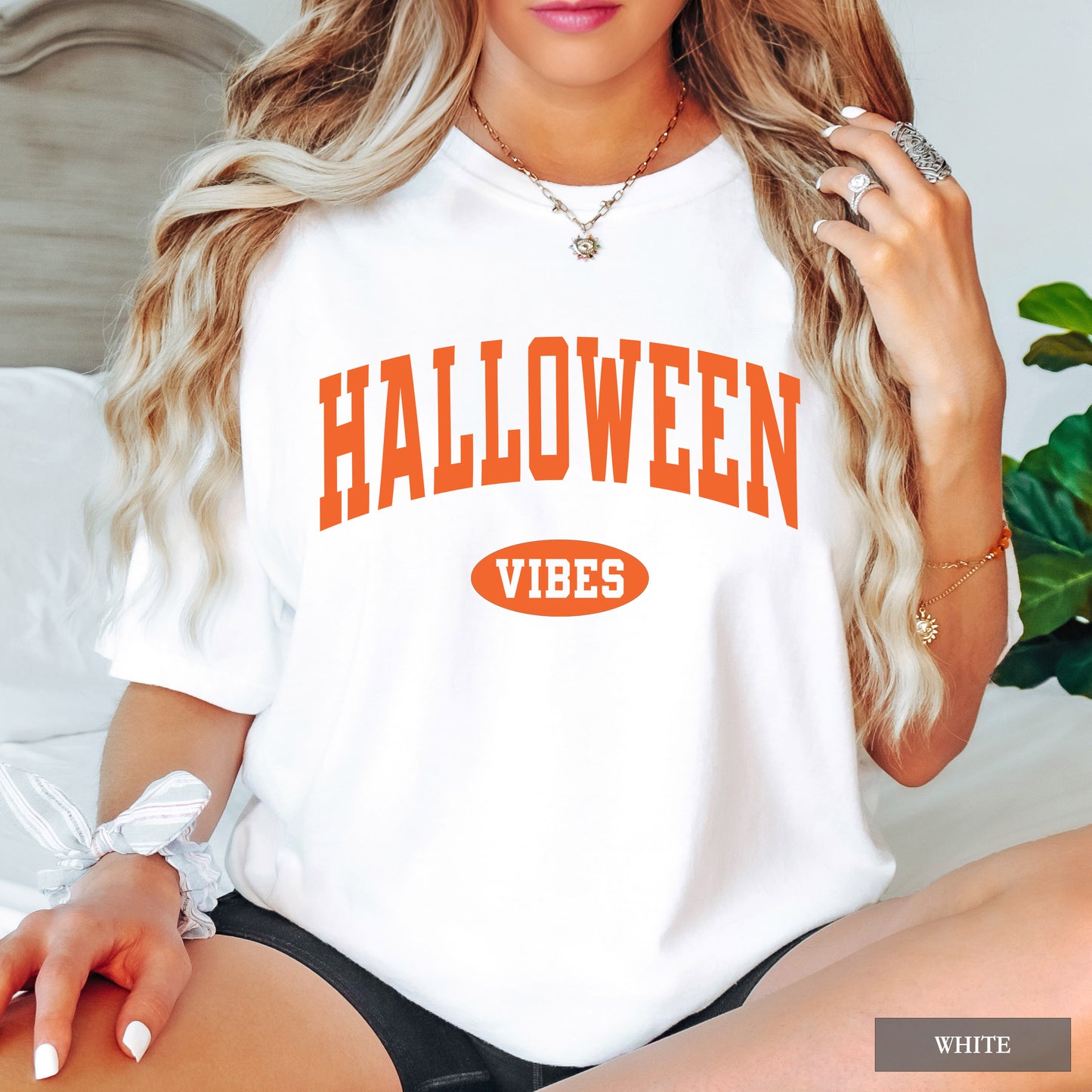A woman wearing a white short-sleeve t-shirt with 'HALLOWEEN VIBES' printed in orange on the front, sitting in a bedroom setting.