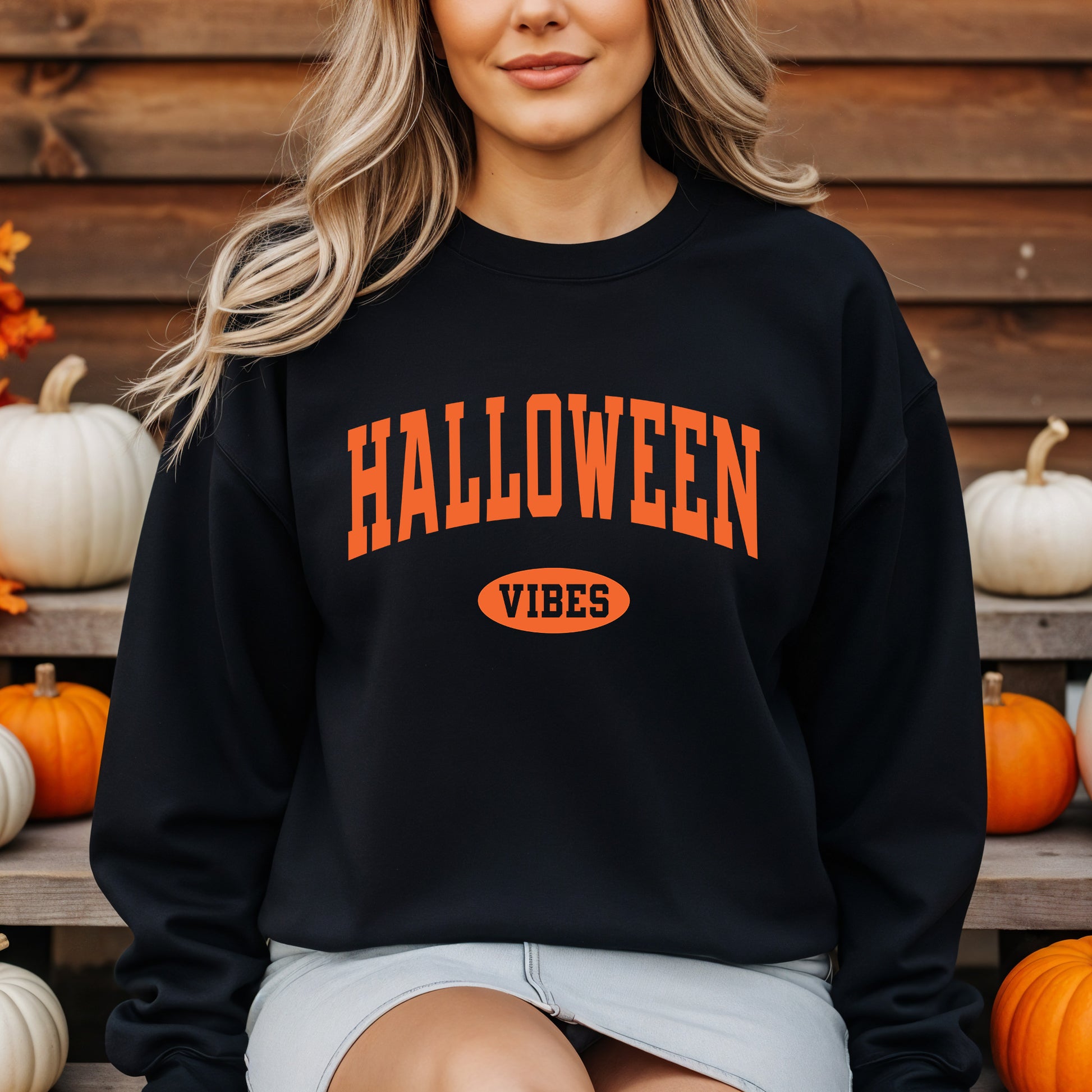 A woman wearing a black crewneck sweatshirt with 'HALLOWEEN VIBES' printed in orange on the front, posed in front of a wooden wall with pumpkins.