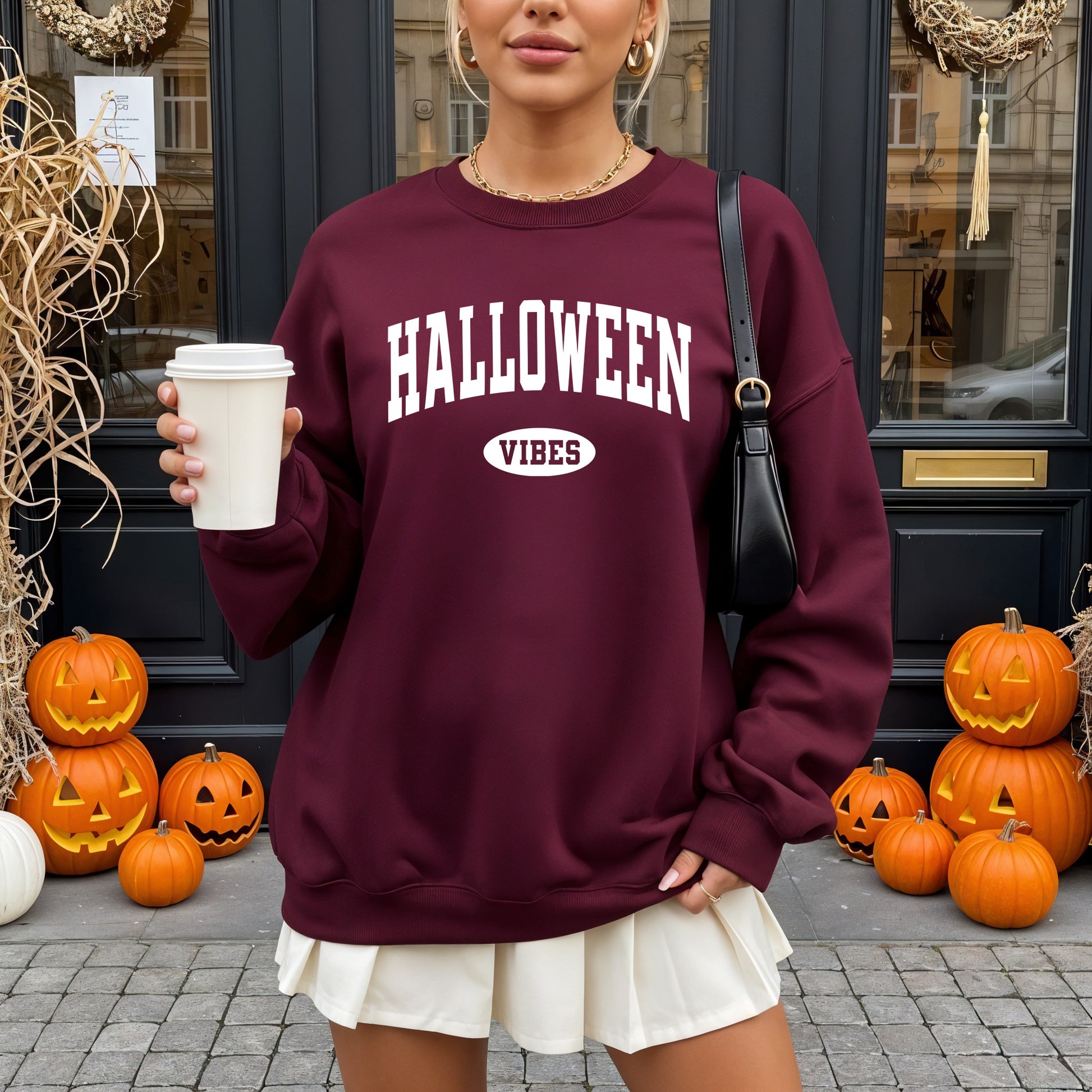 A woman wearing a burgundy sweatshirt with 'HALLOWEEN VIBES' printed in white on the front, holding a coffee cup in front of a Halloween-themed storefront with pumpkins.
