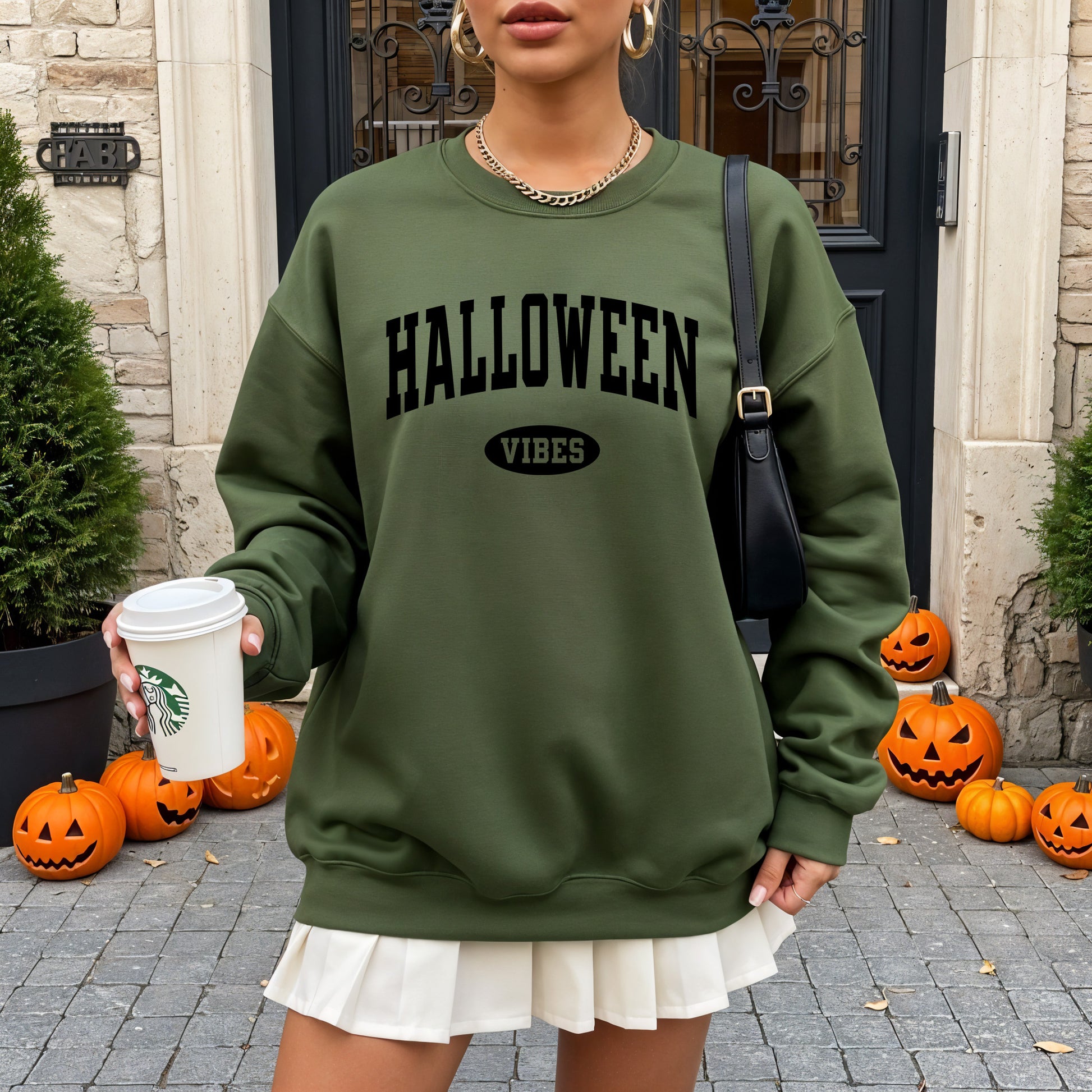 A woman wearing an olive green sweatshirt with 'HALLOWEEN VIBES' printed in black on the front, holding a white coffee cup in front of a stone building with pumpkins around the base.