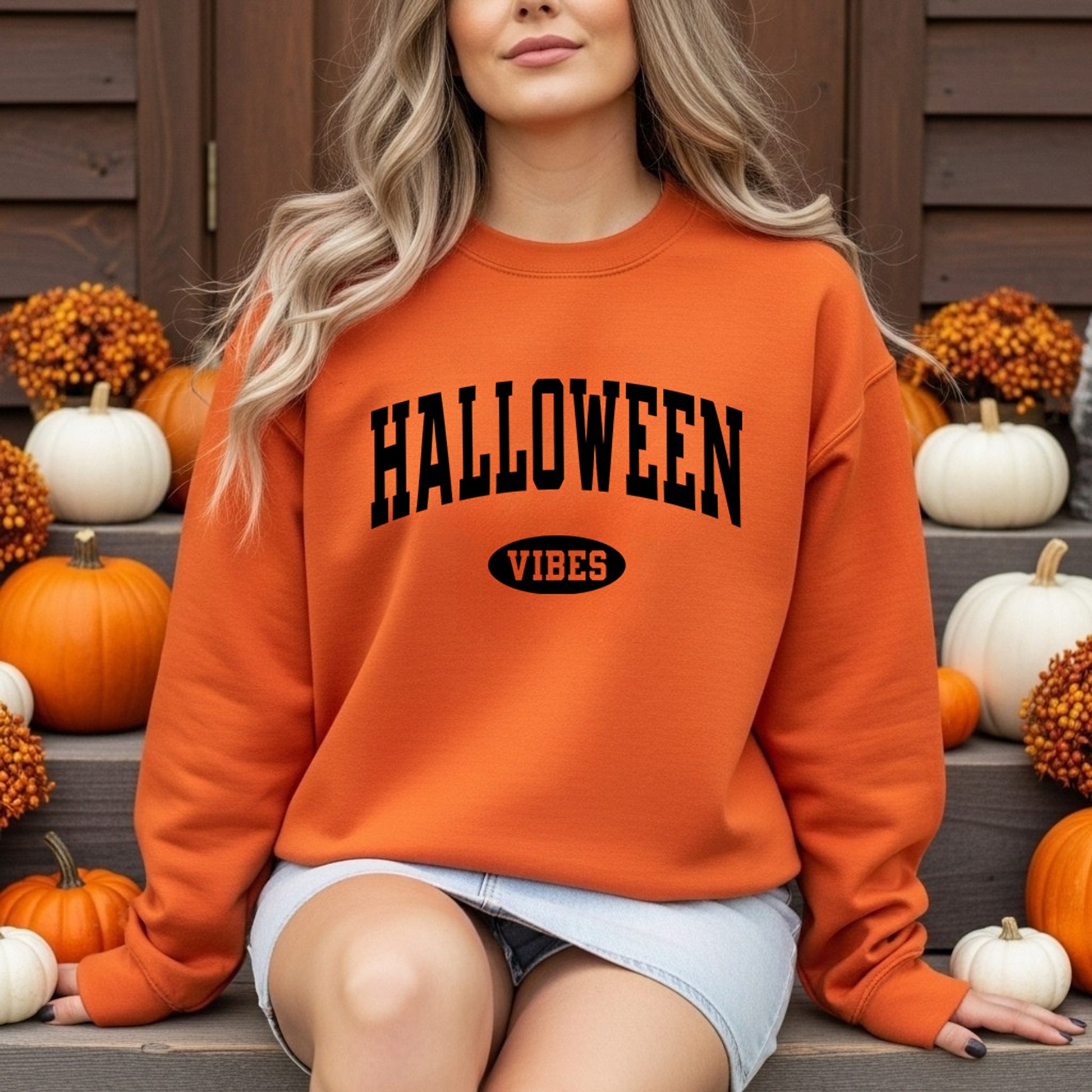 A woman wearing an orange crewneck sweatshirt with 'HALLOWEEN VIBES' printed in black on the front, sitting on steps surrounded by pumpkins and autumn decorations.