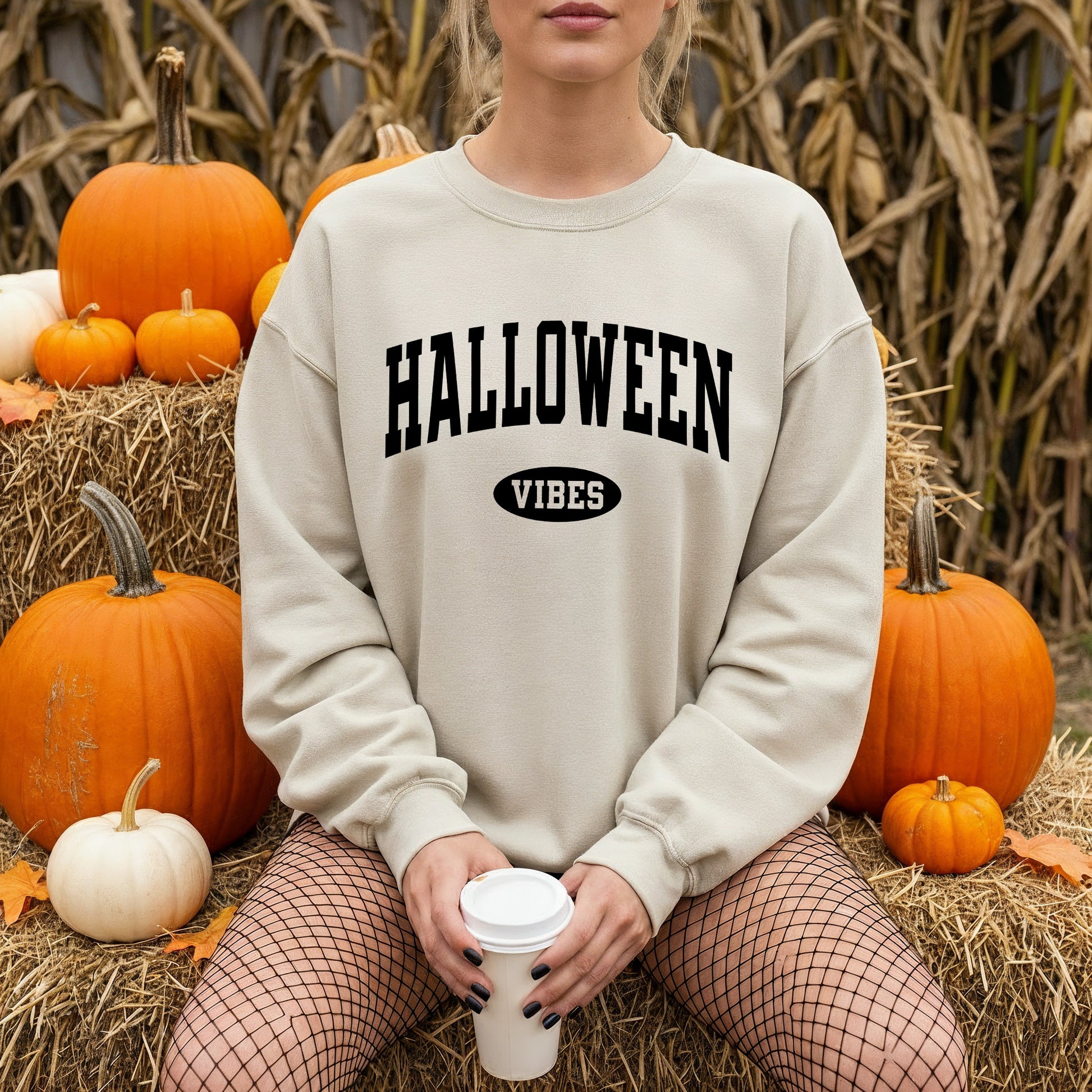 A woman wearing a cream-colored sweatshirt with 'HALLOWEEN VIBES' printed in black on the front, sitting on a hay bale surrounded by pumpkins and corn stalks.