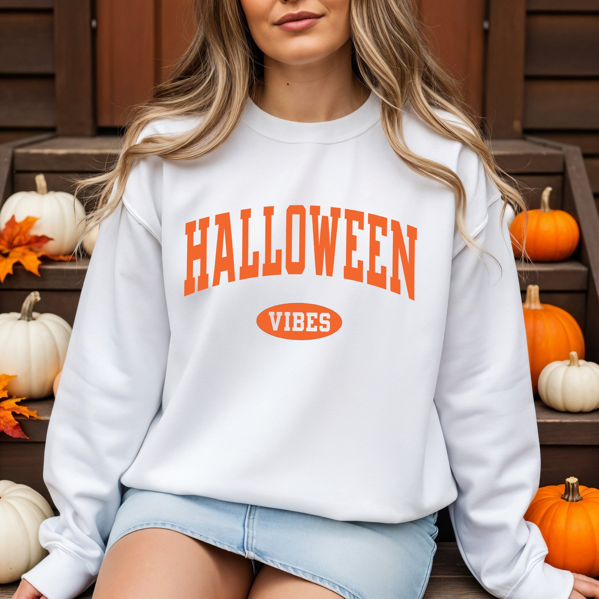 A woman wearing a white crewneck sweatshirt with orange text reading 'HALLOWEEN VIBES' sits on wooden steps decorated with pumpkins and autumn leaves.