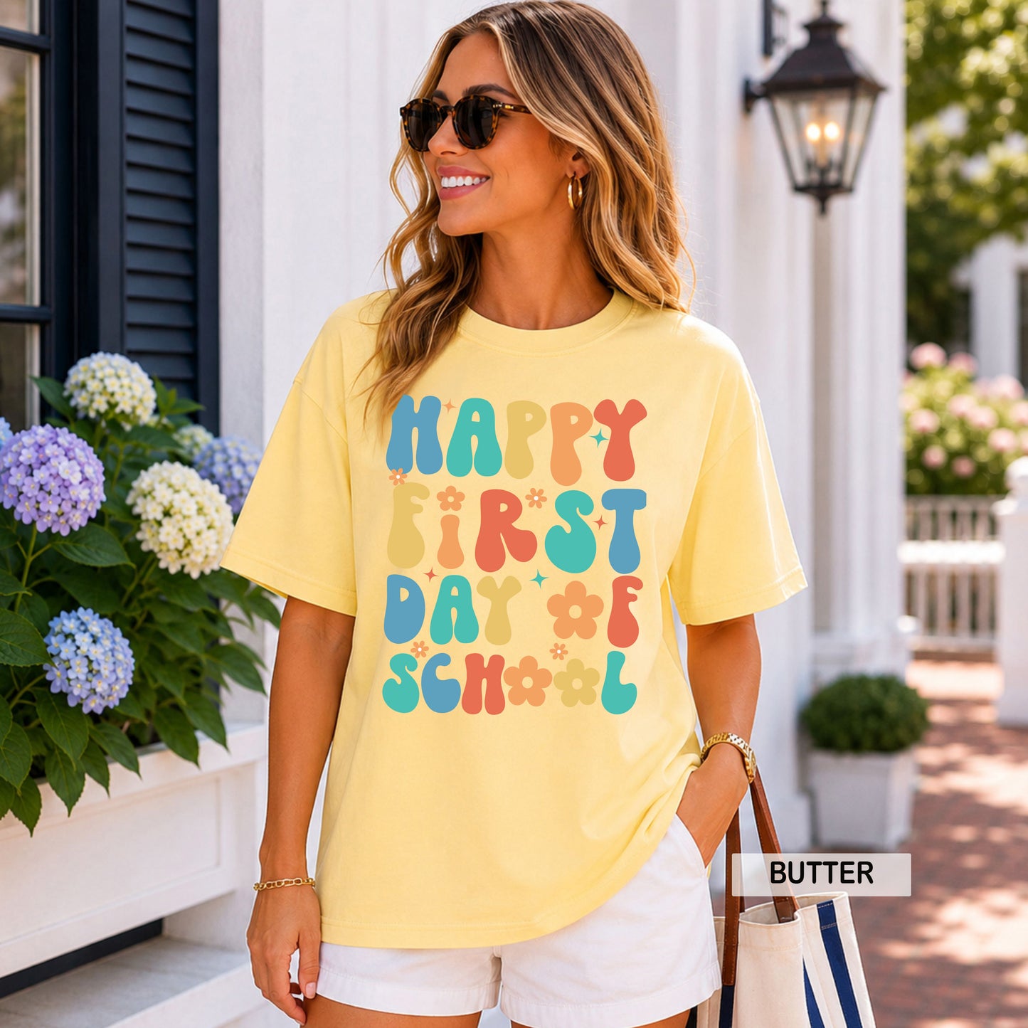 Woman wearing a yellow 'Happy First Day of School' t-shirt in front of a house with flowers.