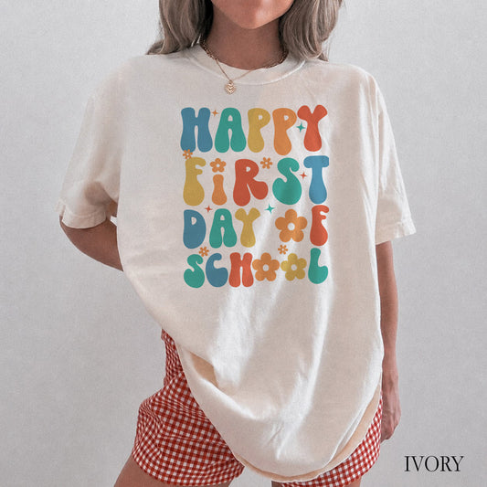 Person wearing a white t-shirt with 'Happy First Day of School' text in colorful letters on a plain background.