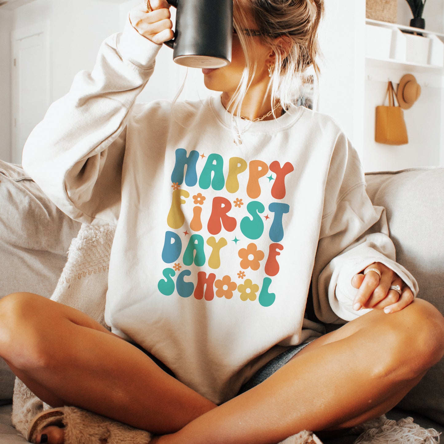 Person wearing a sweatshirt with 'Happy First Day of School' text, holding a mug.