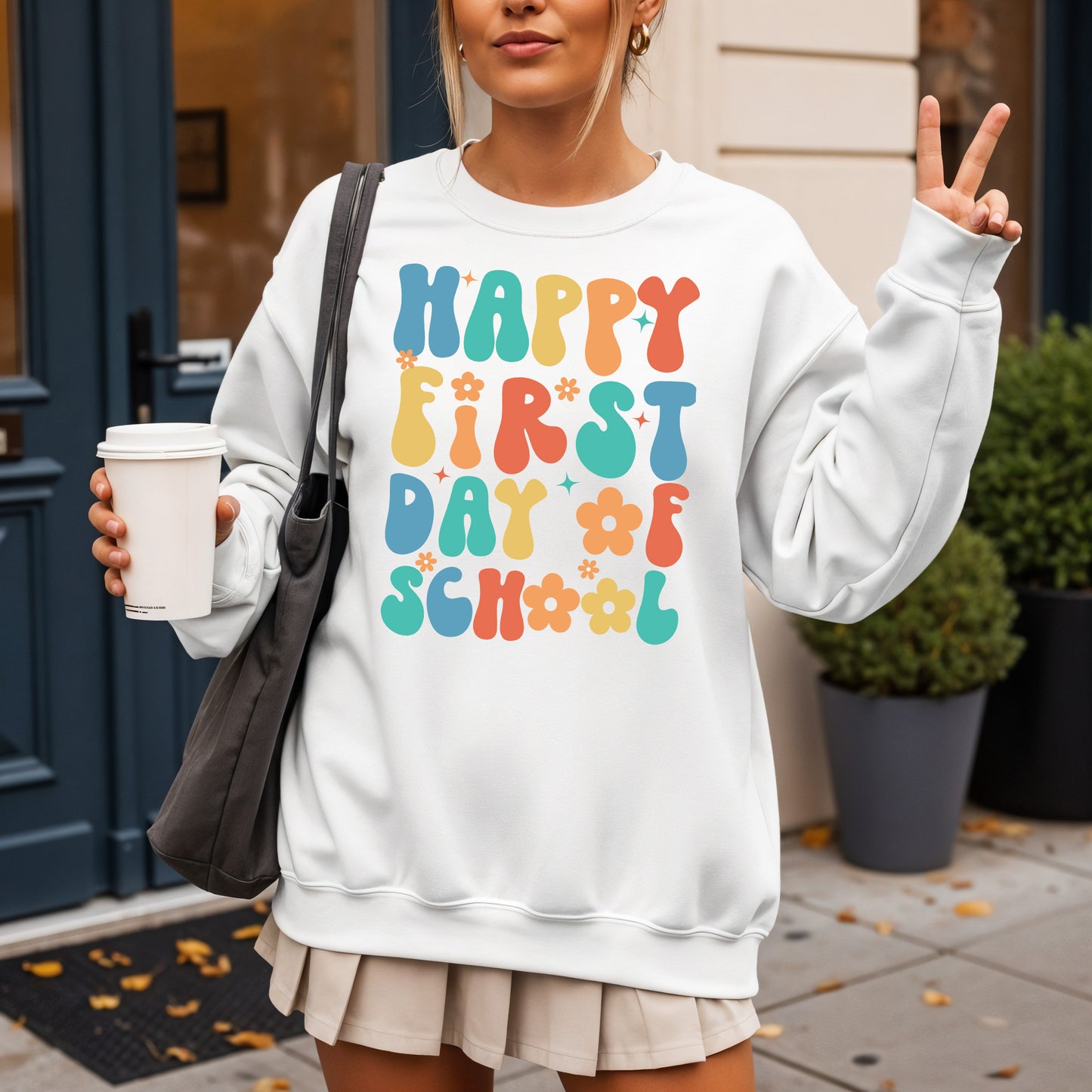Person wearing a white sweatshirt with colorful 'Happy First Day of School' text, holding a coffee cup.
