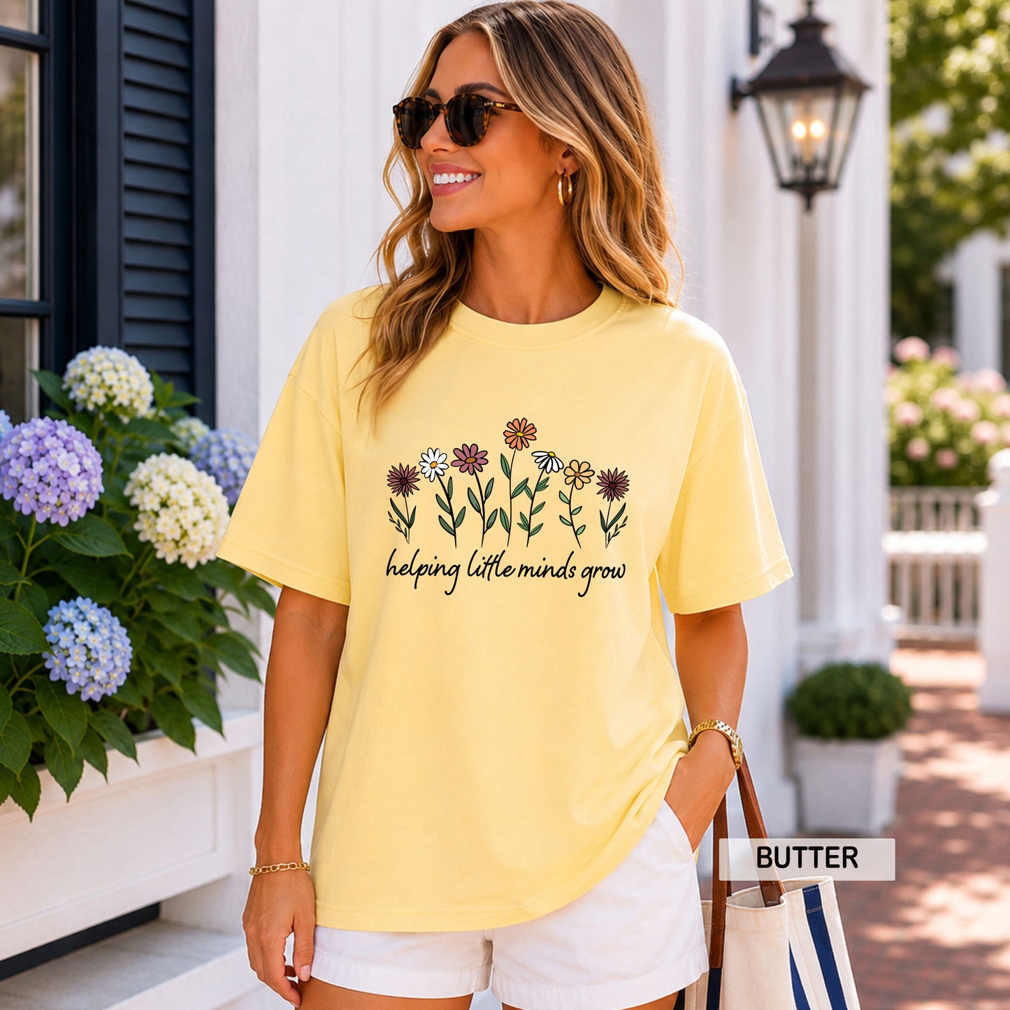 Woman wearing a yellow 'TEACHER' t-shirt with colorful flowers in front of a white building.