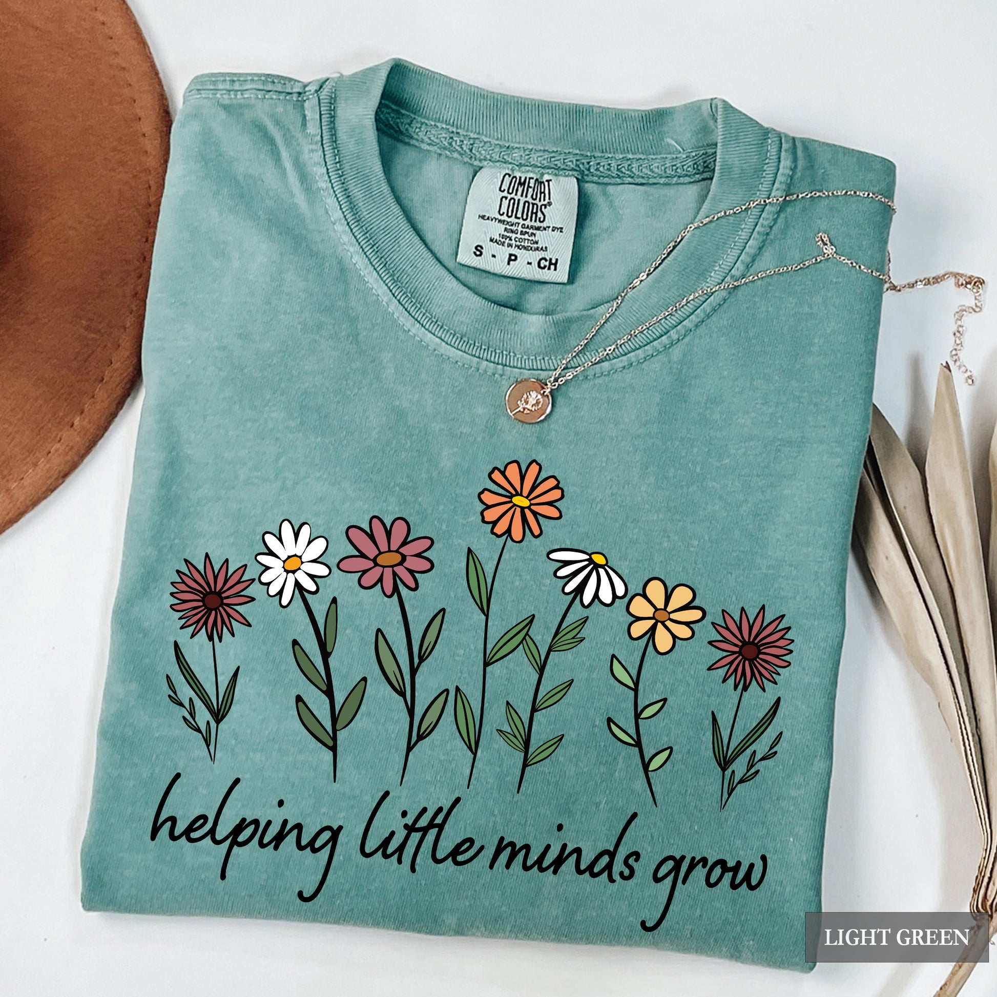 Light green t-shirt with floral design and text 'helping little minds grow' on a white background.