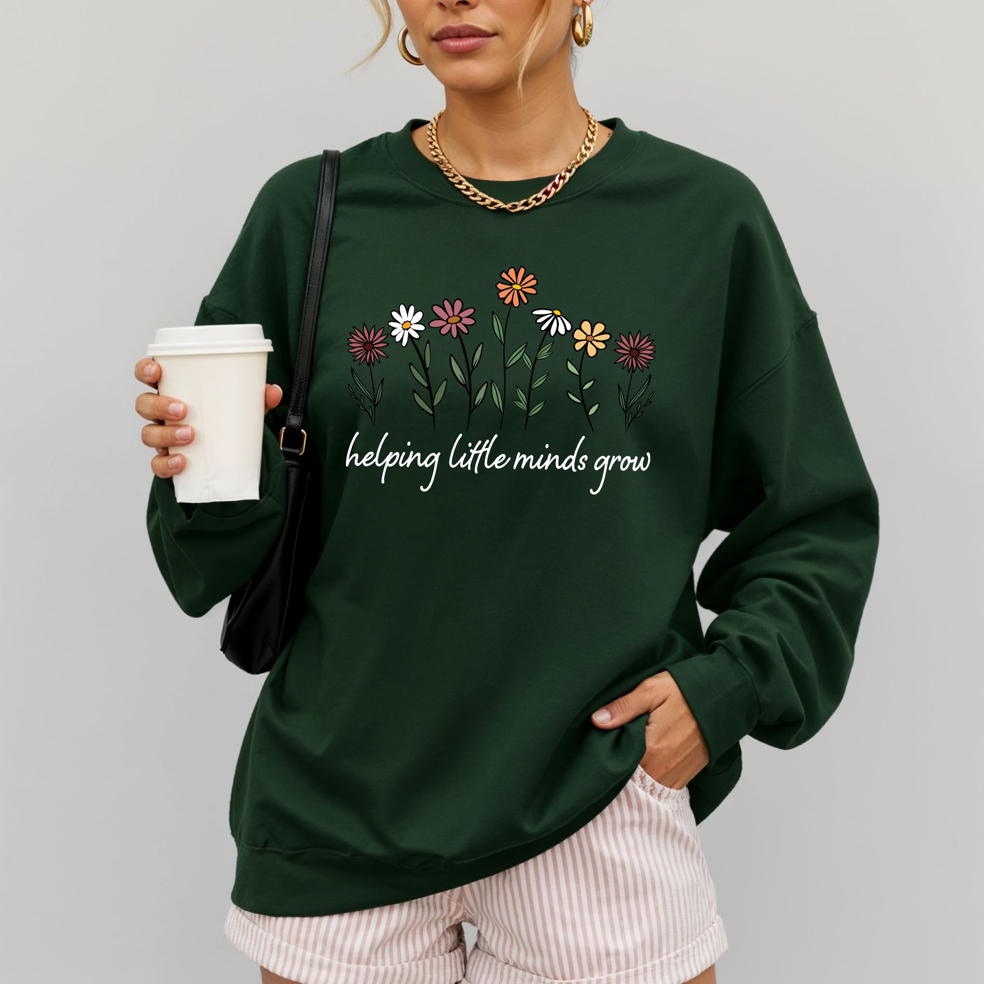 Person wearing a green sweatshirt with floral design and text, holding a coffee cup.