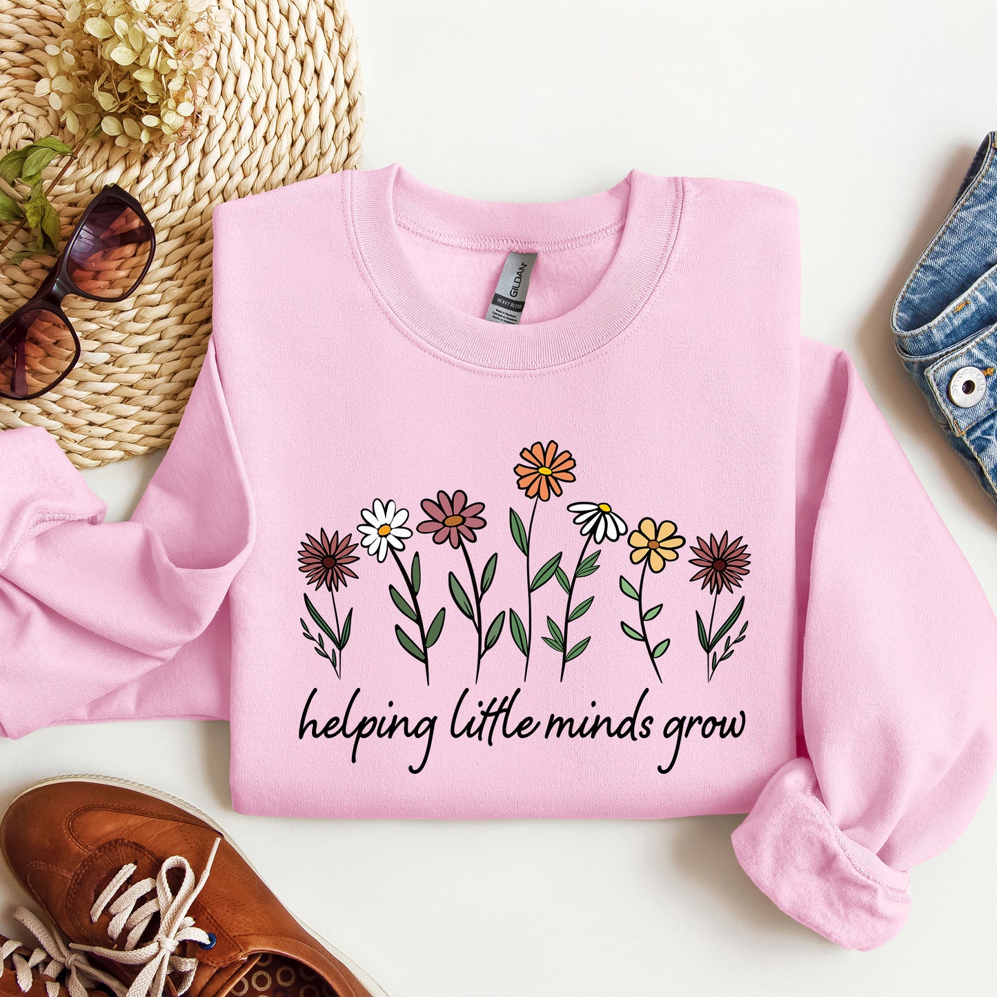 Pink sweatshirt with floral design and text 'helping little minds grow' on a white surface with accessories.