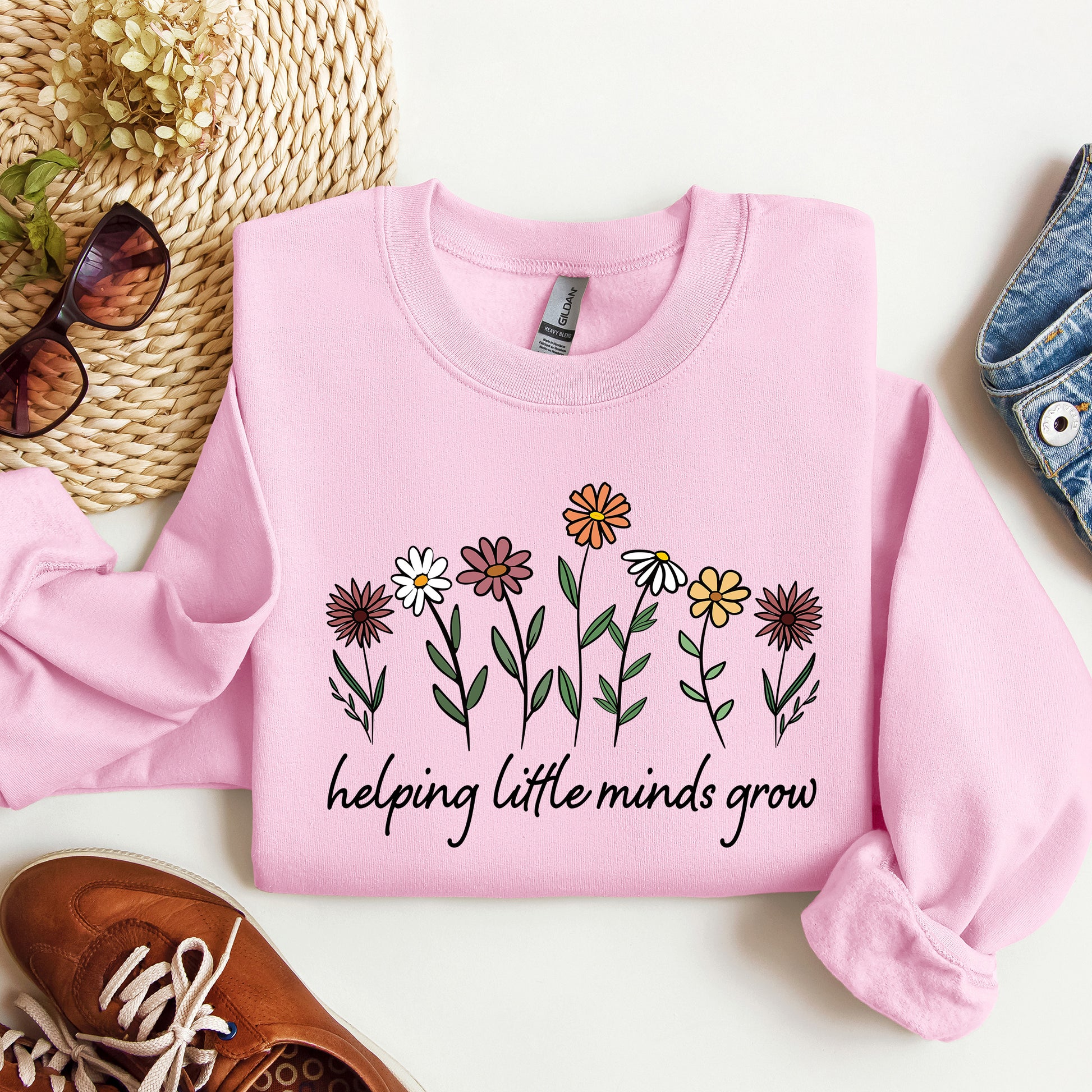 Pink sweatshirt with floral design and text 'helping little minds grow' on a white surface with accessories.