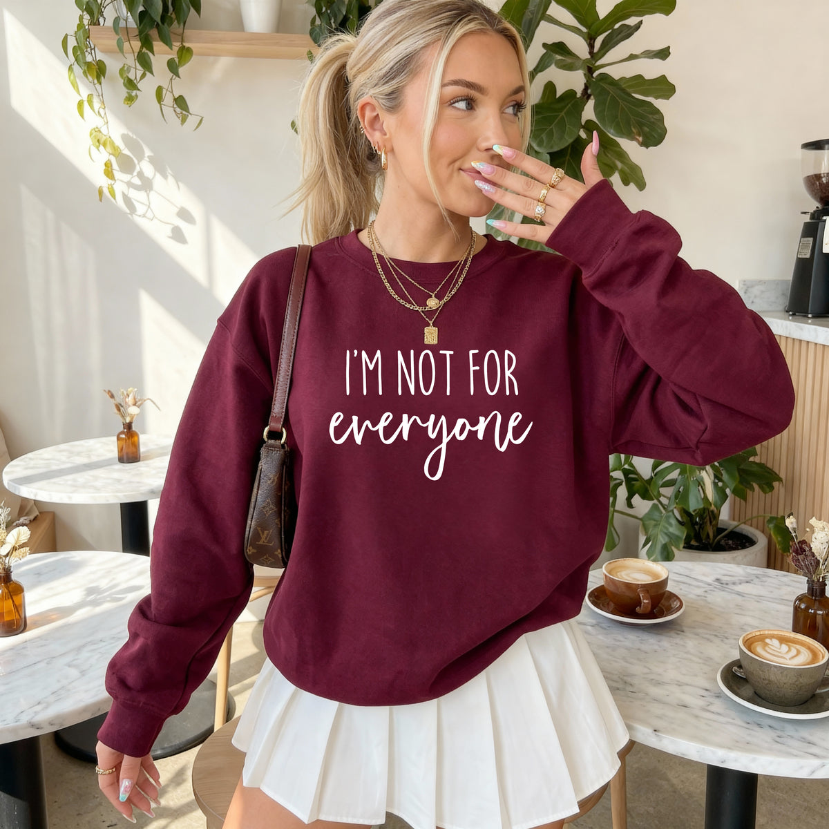 A woman in a cafe setting wearing a maroon sweatshirt with the white text 'I'M NOT FOR everyone' and a white pleated skirt.