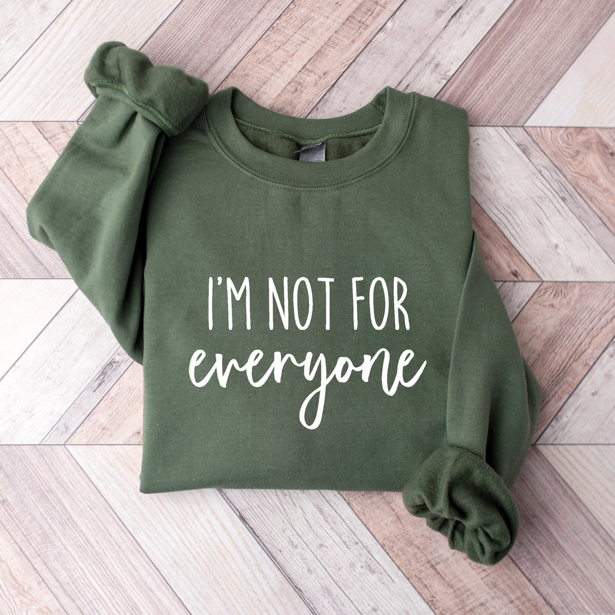 An olive green crewneck sweatshirt with the text 'I'M NOT FOR everyone' printed in white on the front, laid flat on a light wood chevron-patterned floor.